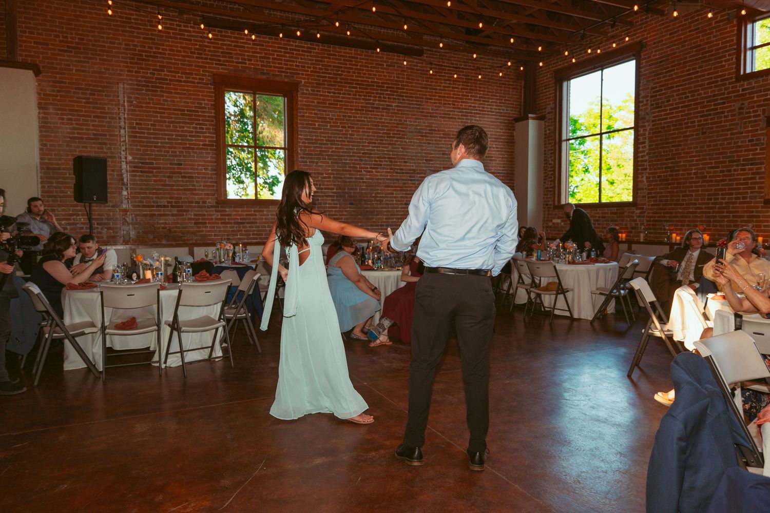 Wedding guests dancing and celebrating on the dance floor of a rustic brick venue with string lights overhead.