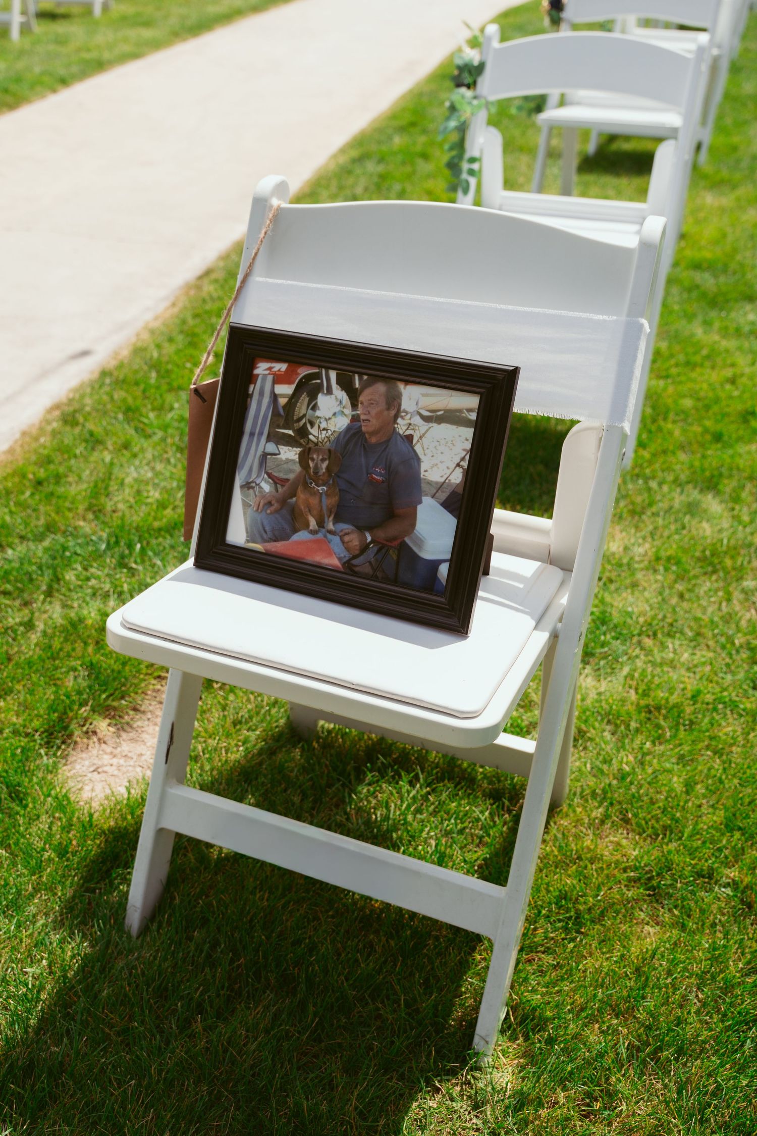 White folding chairs arranged on grass with small picture frames attached to their backs.