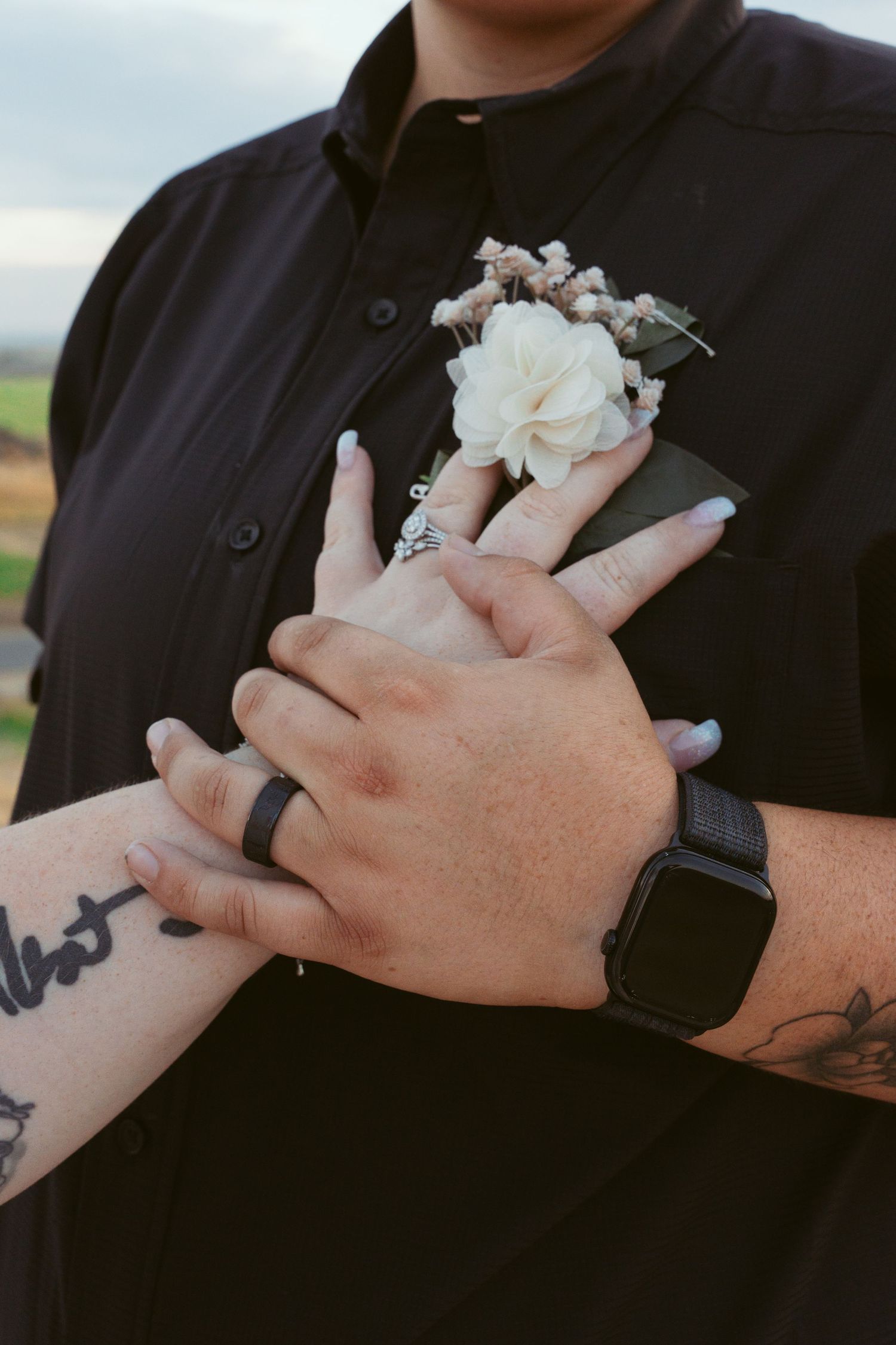 Close-up of hands wearing wedding rings with white boutonniere and visible tattoo details.