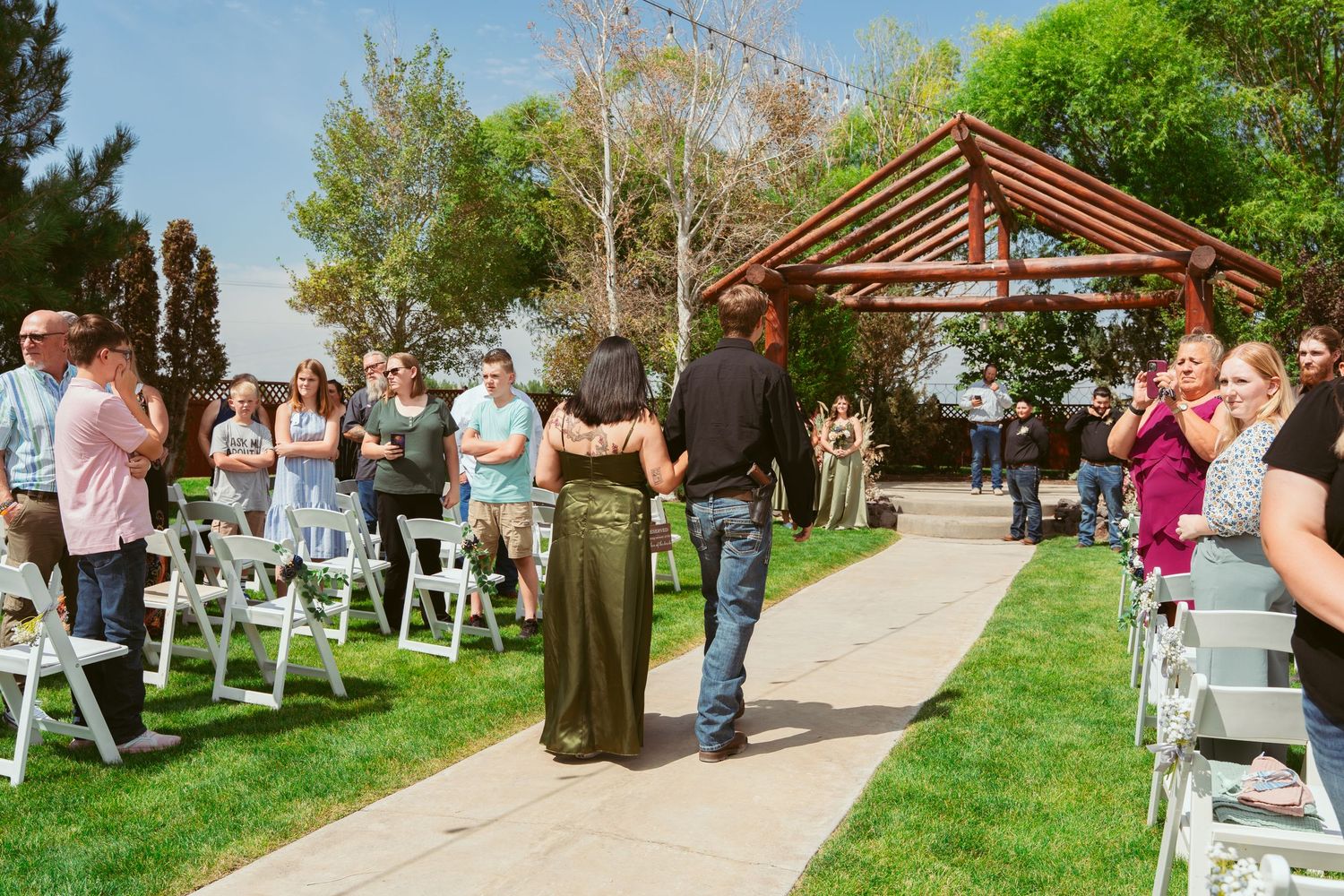 Guests seated in white chairs observe an outdoor wedding ceremony under bright blue skies and green trees.