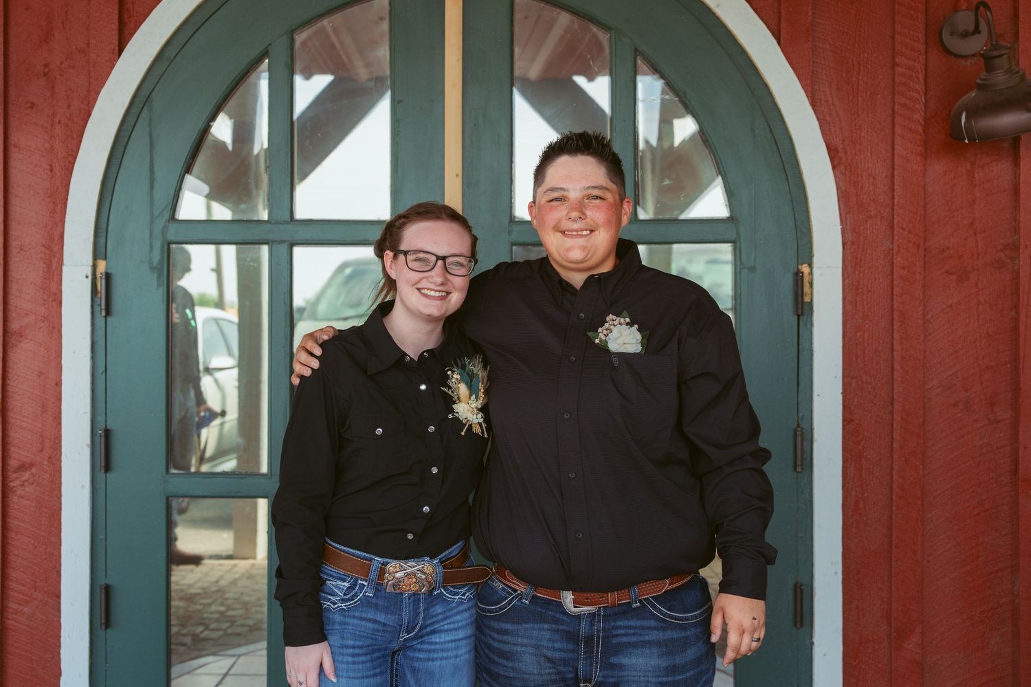 Two people stand together smiling in front of a red building with teal arched doorway.