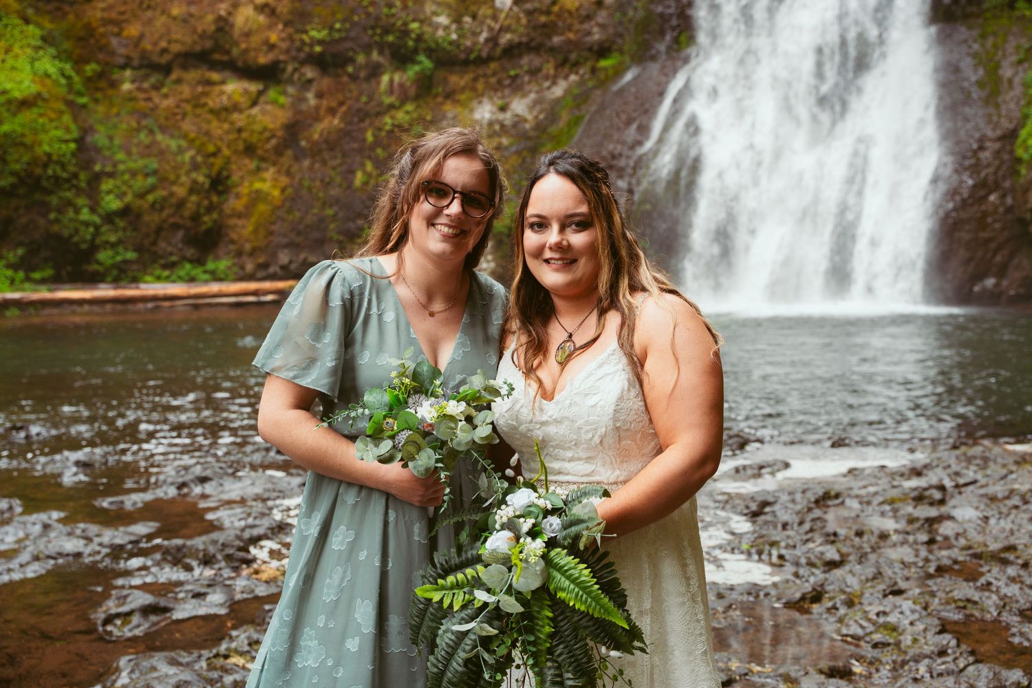 Wedding party members gather near a scenic waterfall holding fresh green and white bouquets.