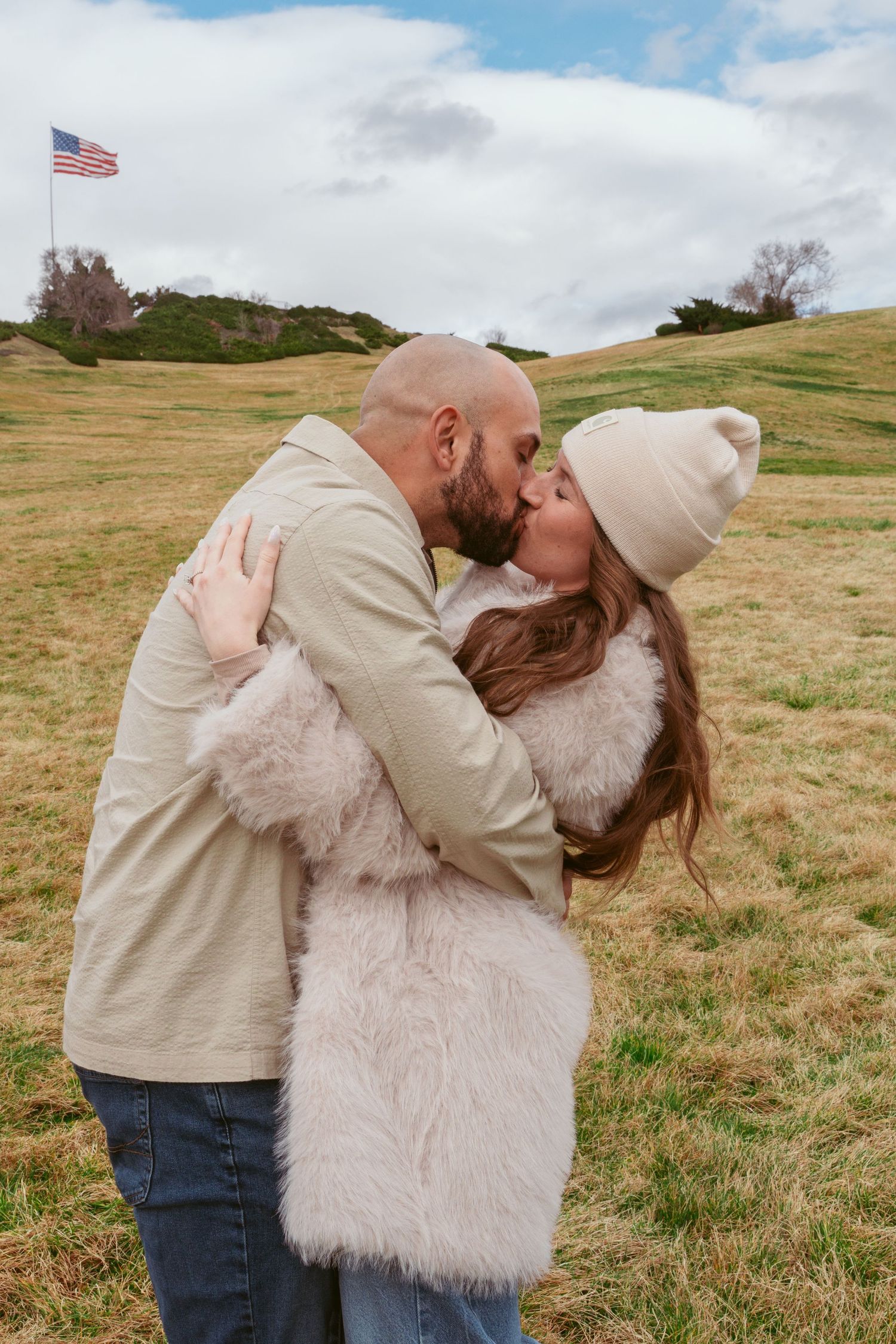 A couple embraces in an open field with American flags visible in the distance.