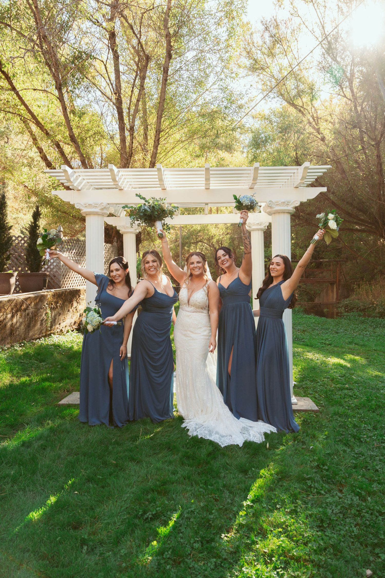 Wedding party in dusty blue dresses poses under white pergola in garden setting.