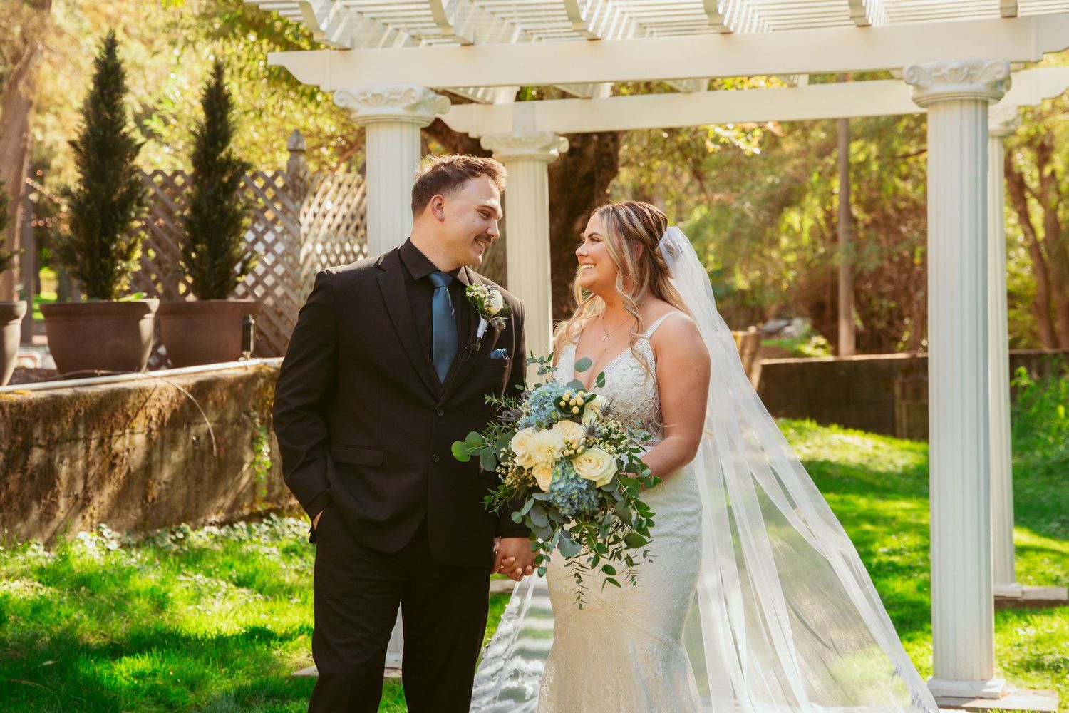 A wedding couple shares a romantic moment under a white pergola in a garden setting.