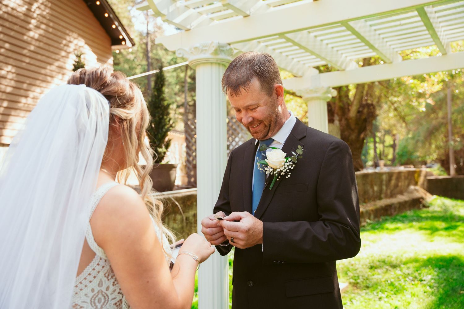 Newlyweds exchange vows in a garden setting beneath a white pergola on a sunny day.