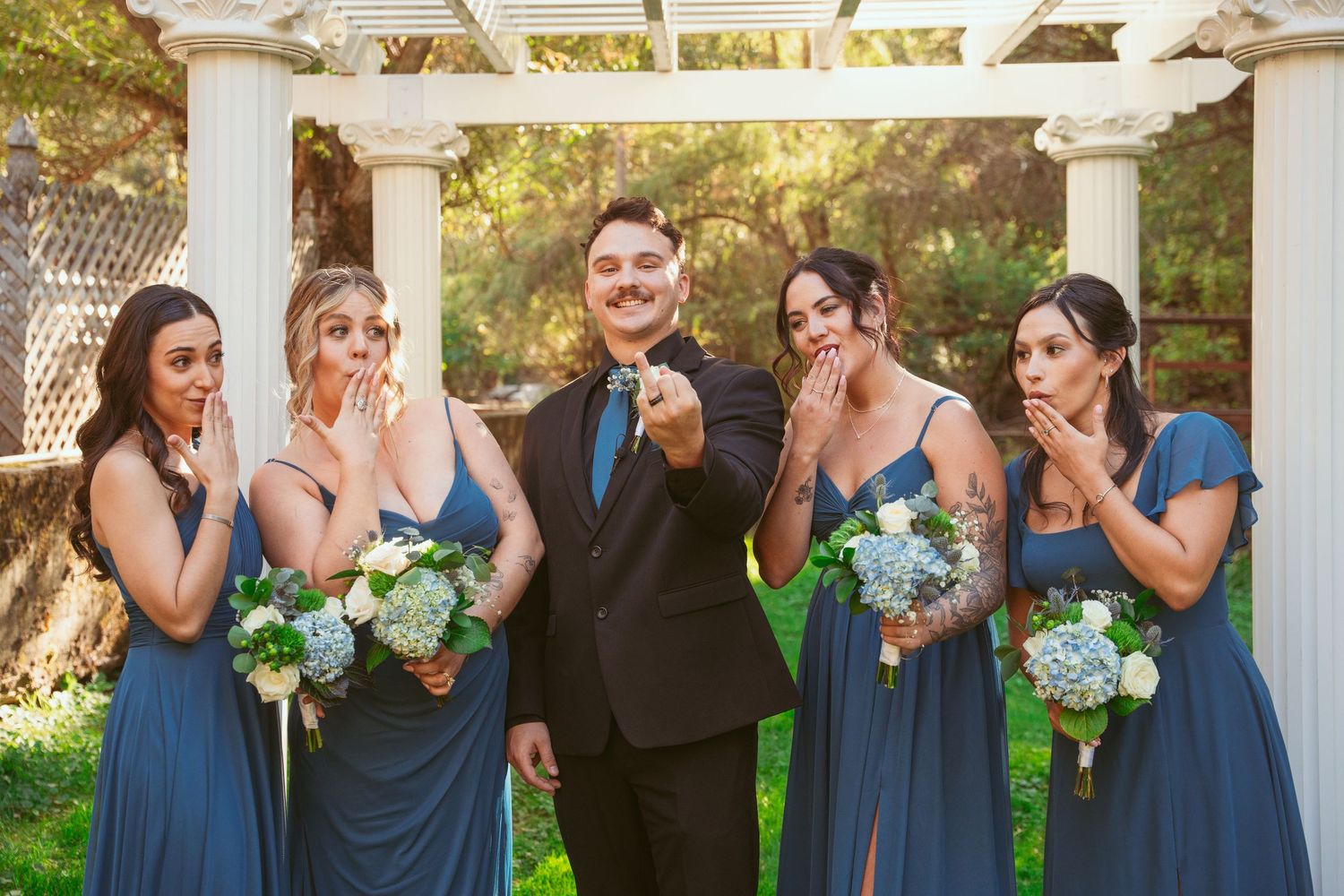 Wedding party in blue dresses and black suits pose together under a white pergola.