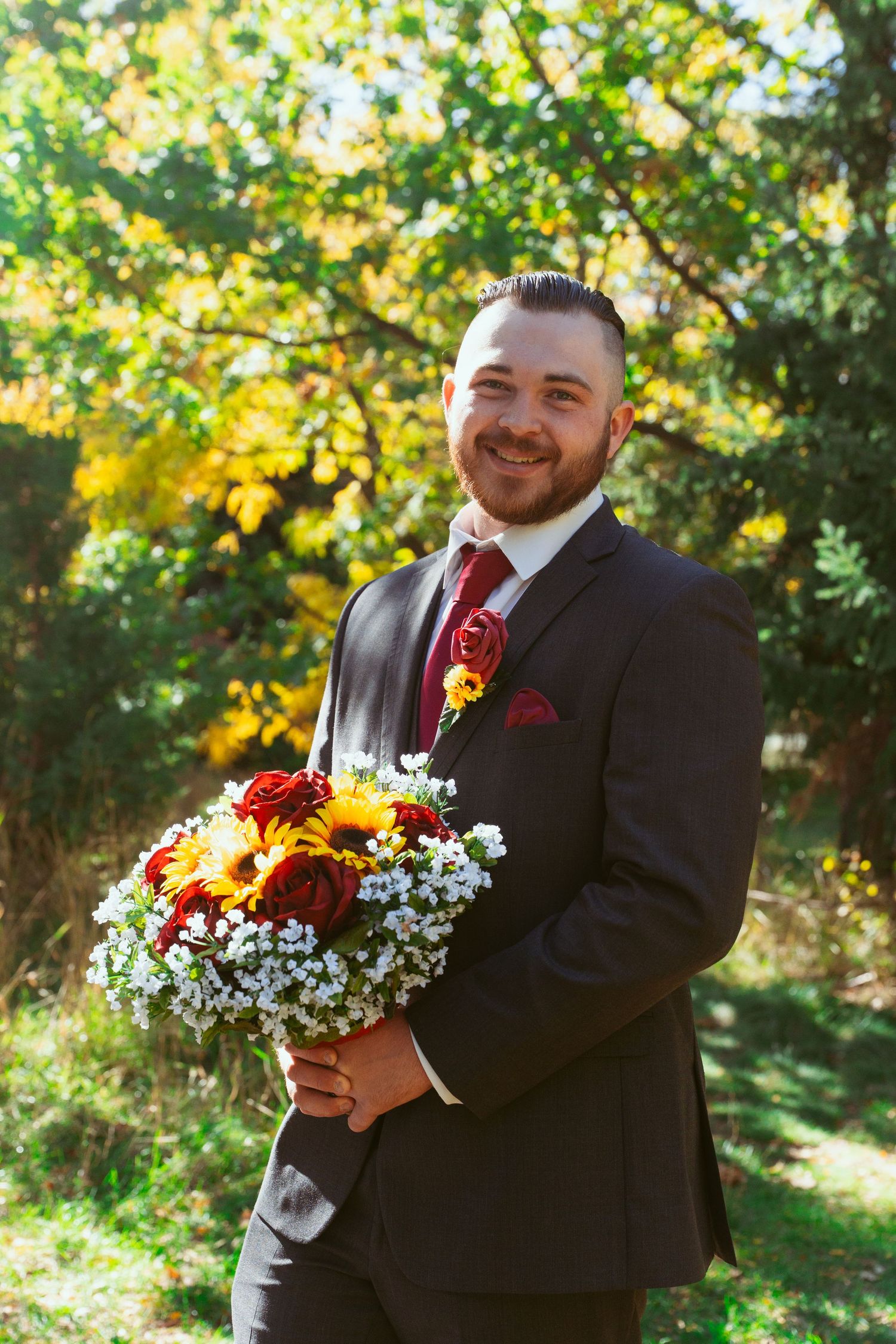 Wedding party members in dark suits holding bright sunflower and daisy bouquets against autumn foliage.
