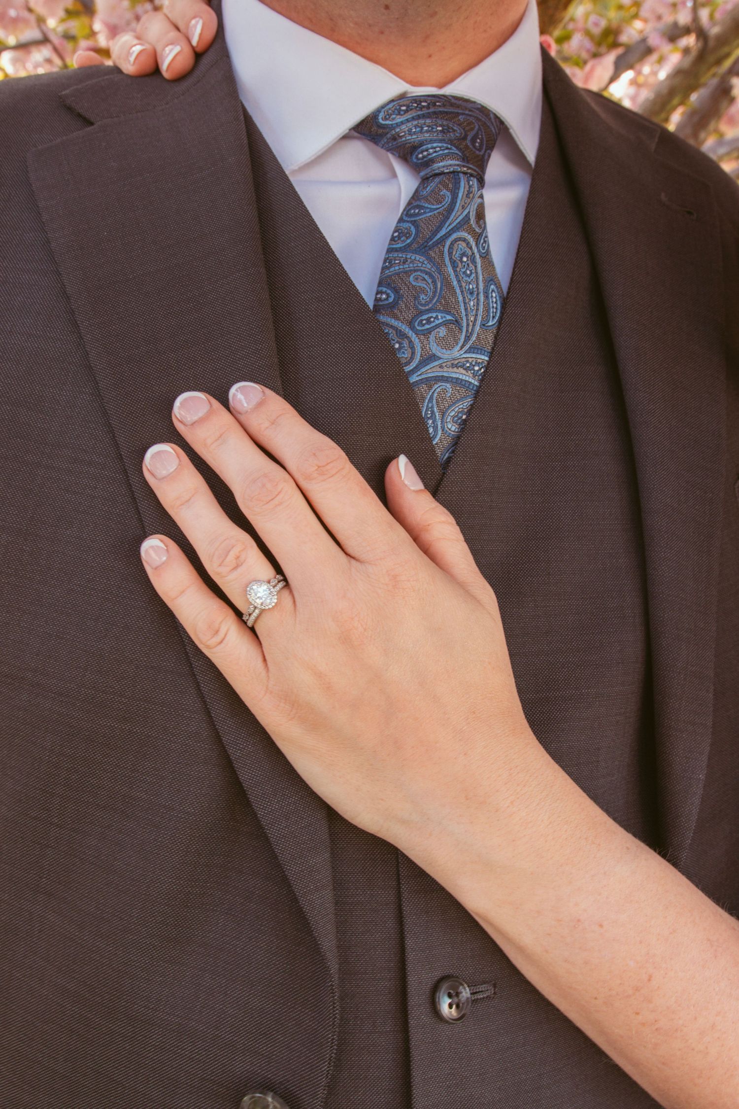 A hand wearing a wedding ring rests on a brown suit lapel with a light blue shirt and patterned blue tie visible.