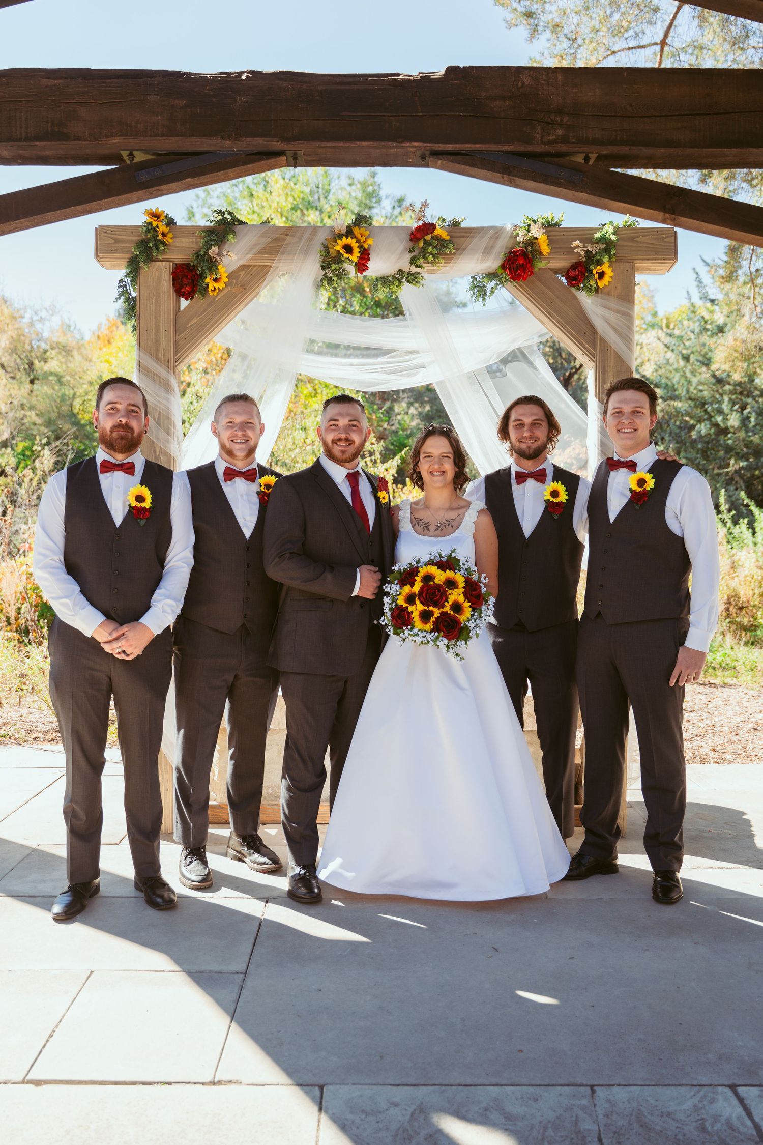 Wedding party in brown vests and formal attire poses under a decorated outdoor pavilion with sunflowers and white fabric.