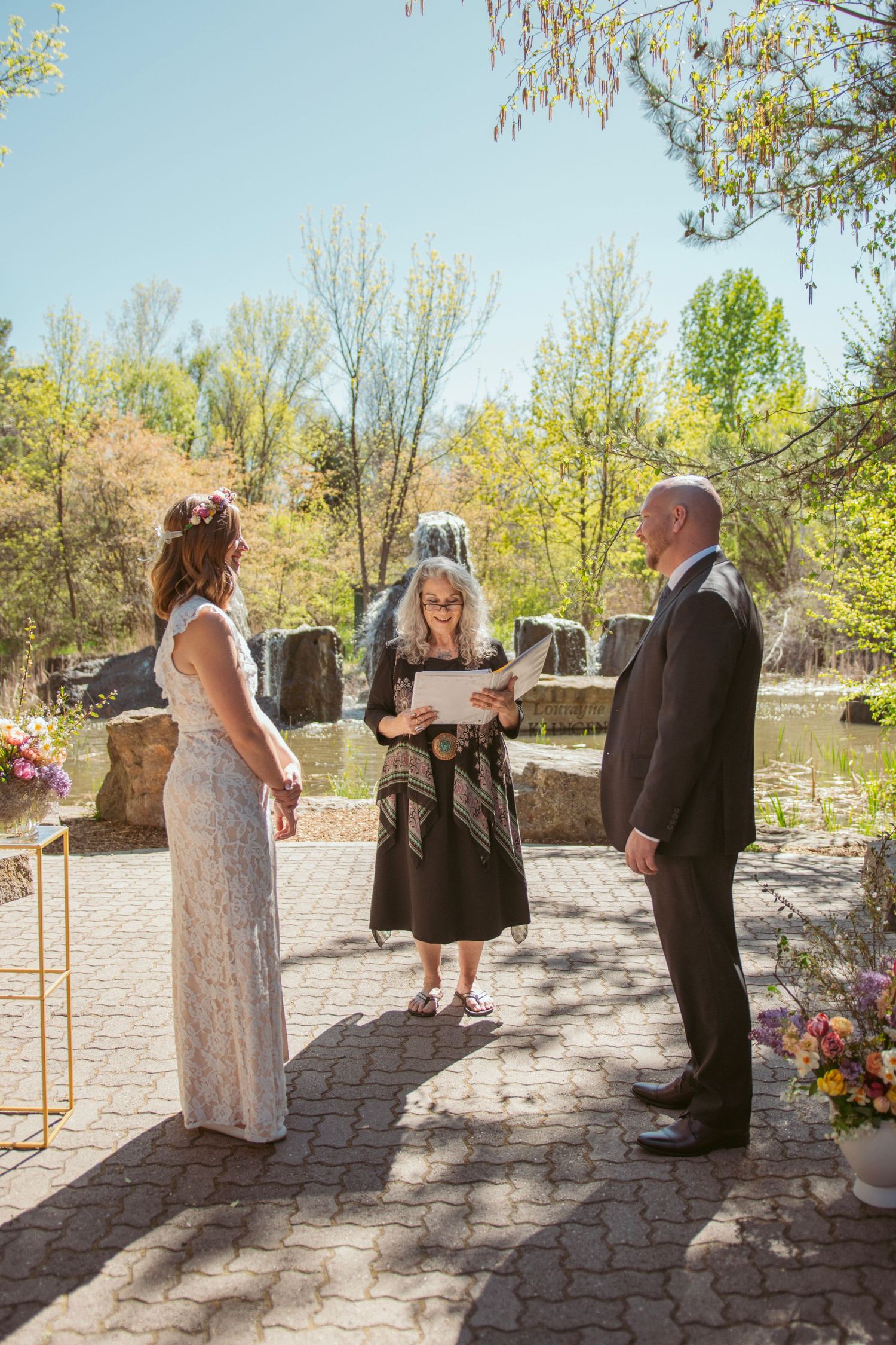 Outdoor wedding ceremony taking place on a stone patio with trees and flowers in the background.