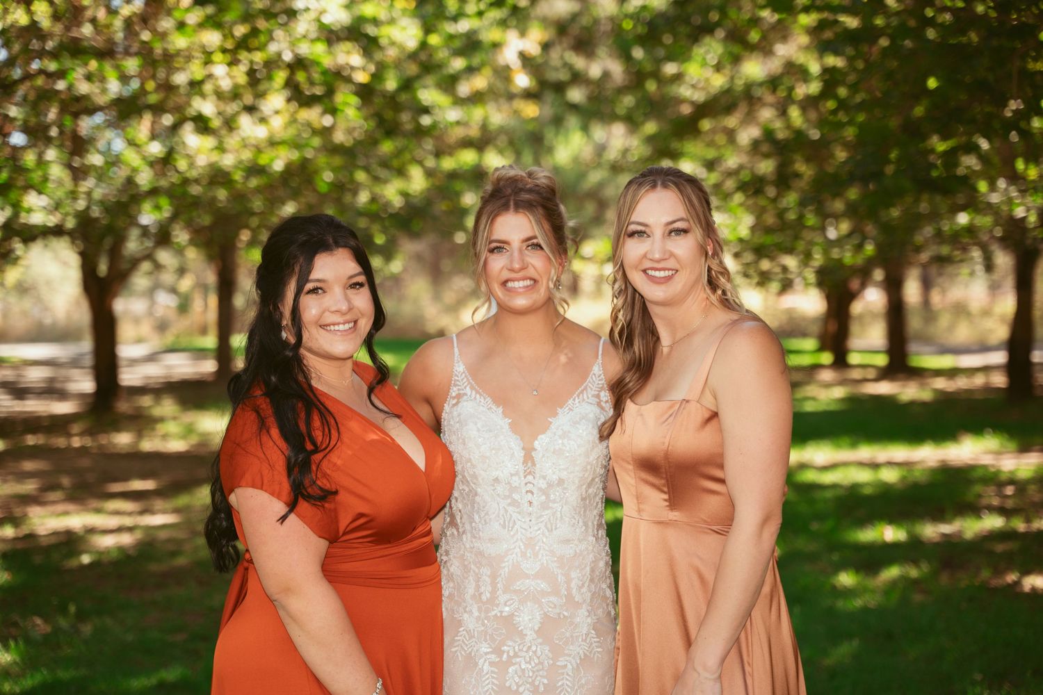 Bridal party in orange dresses and white gown gather for photos in a green outdoor setting.