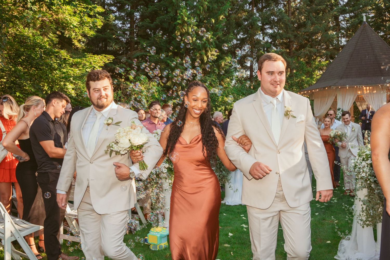 Wedding party in cream suits and coral dresses poses on lawn during outdoor celebration.