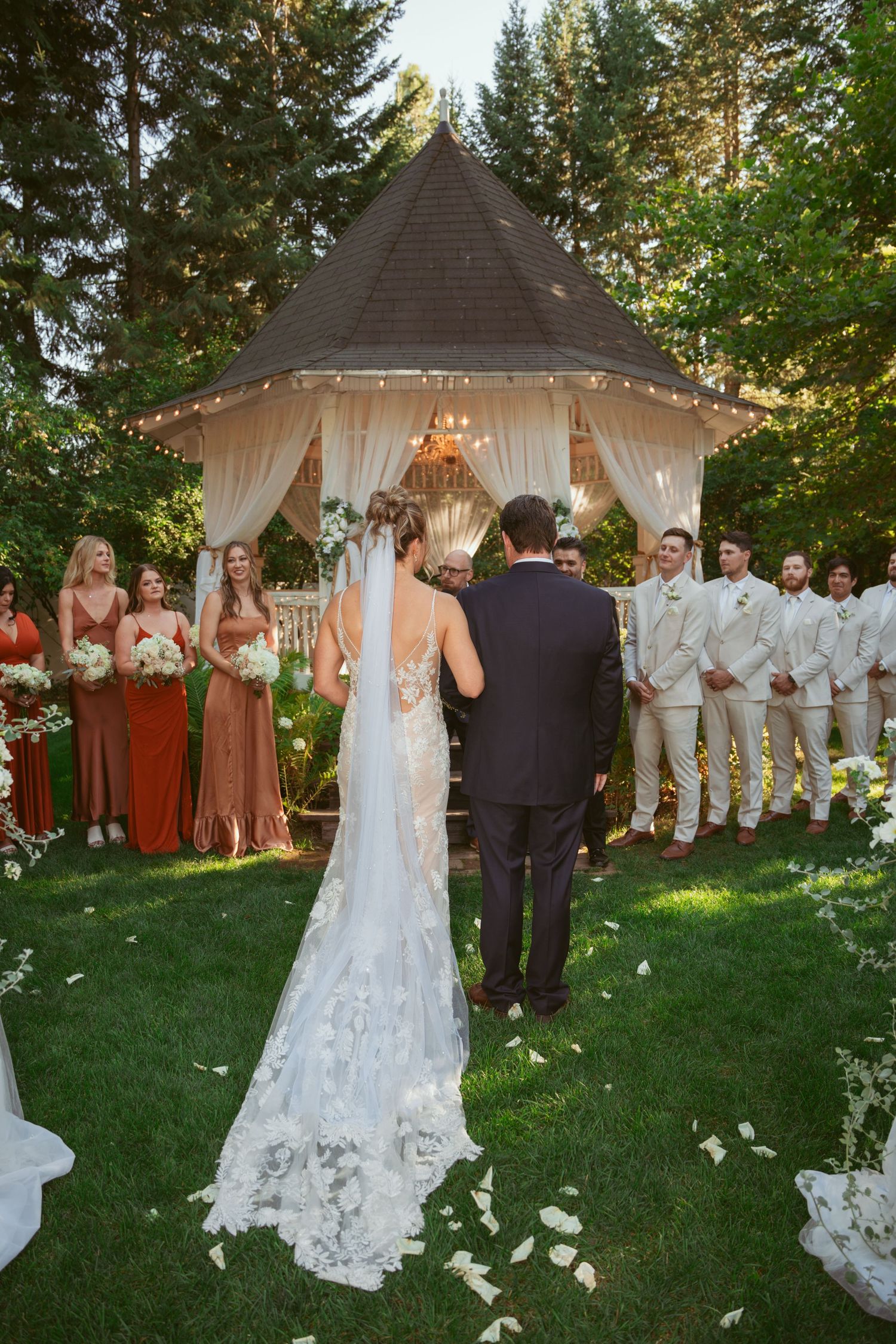 A wedding ceremony takes place under a gazebo with white drapes, surrounded by bridesmaids in rust dresses and groomsmen in beige.