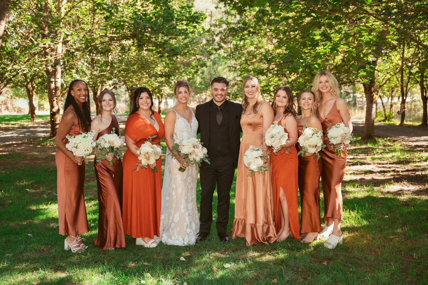 Wedding party in rust-colored dresses and white gowns poses together in a garden setting.