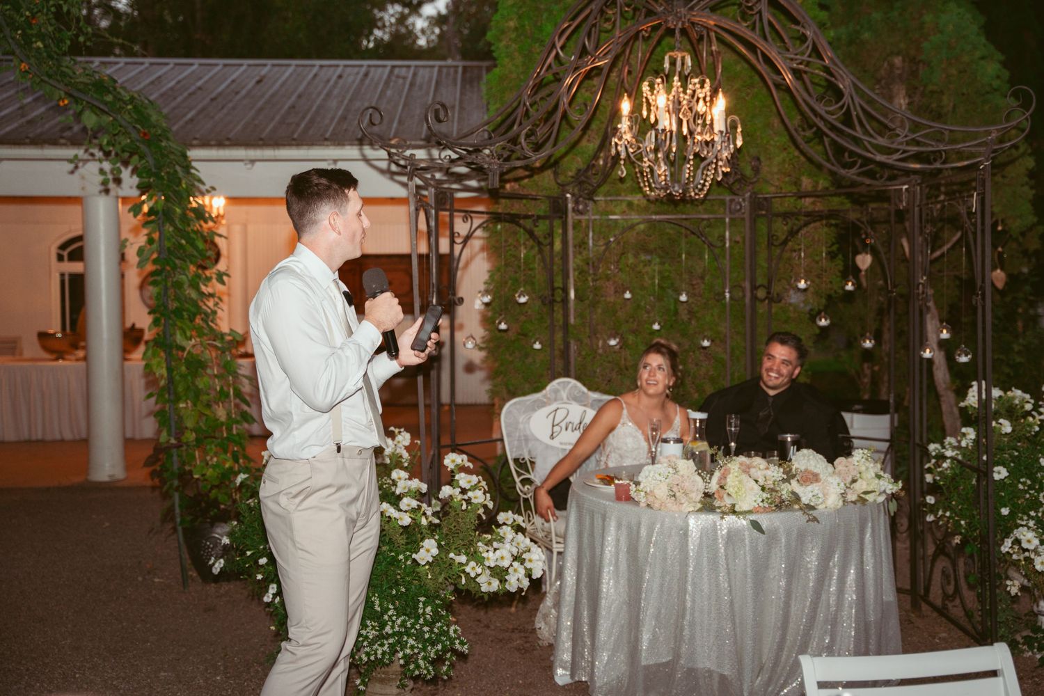 A wedding speech being delivered under an elegant floral arch with a crystal chandelier at an outdoor reception.