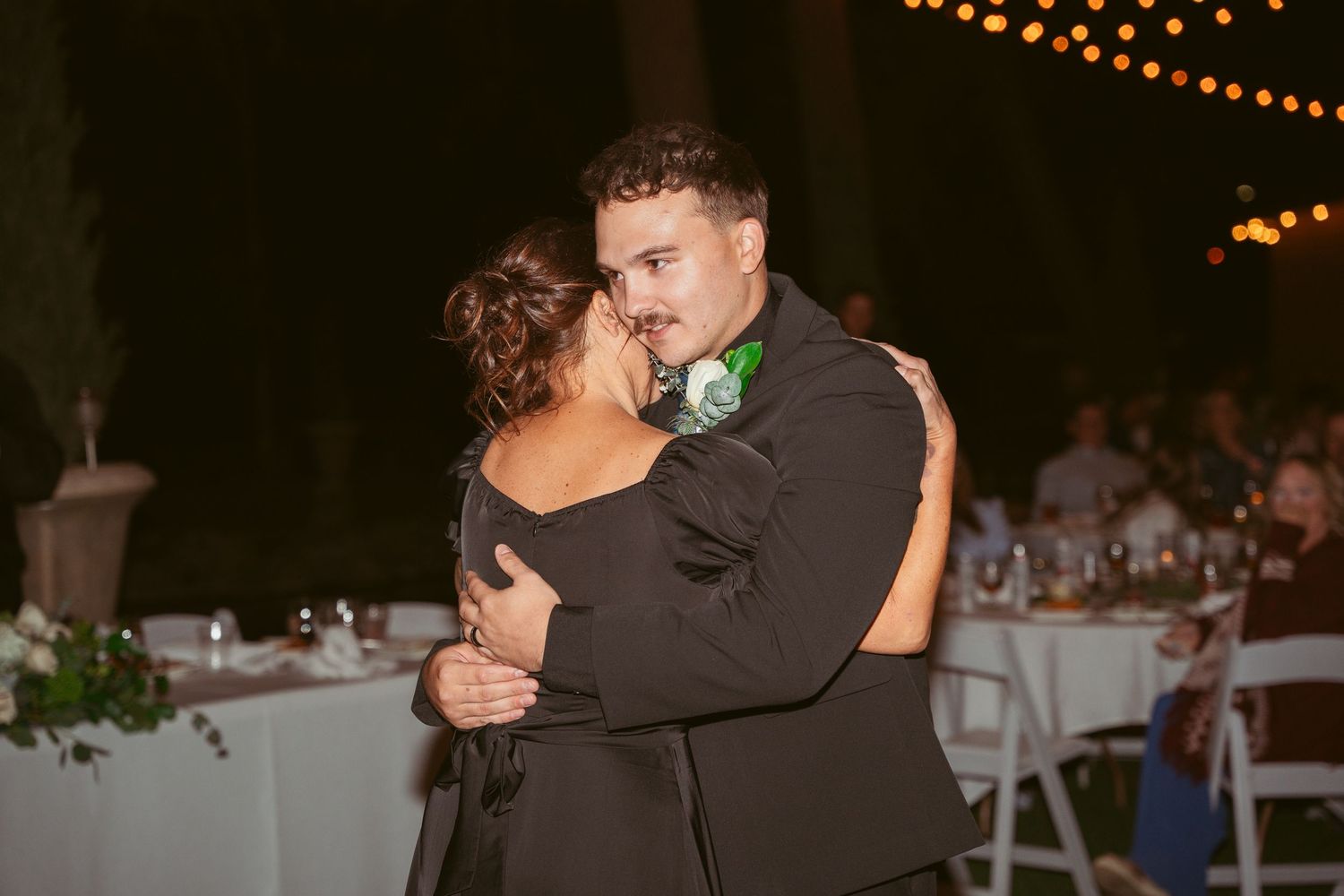 A couple shares a romantic slow dance at an outdoor evening wedding reception with string lights overhead.