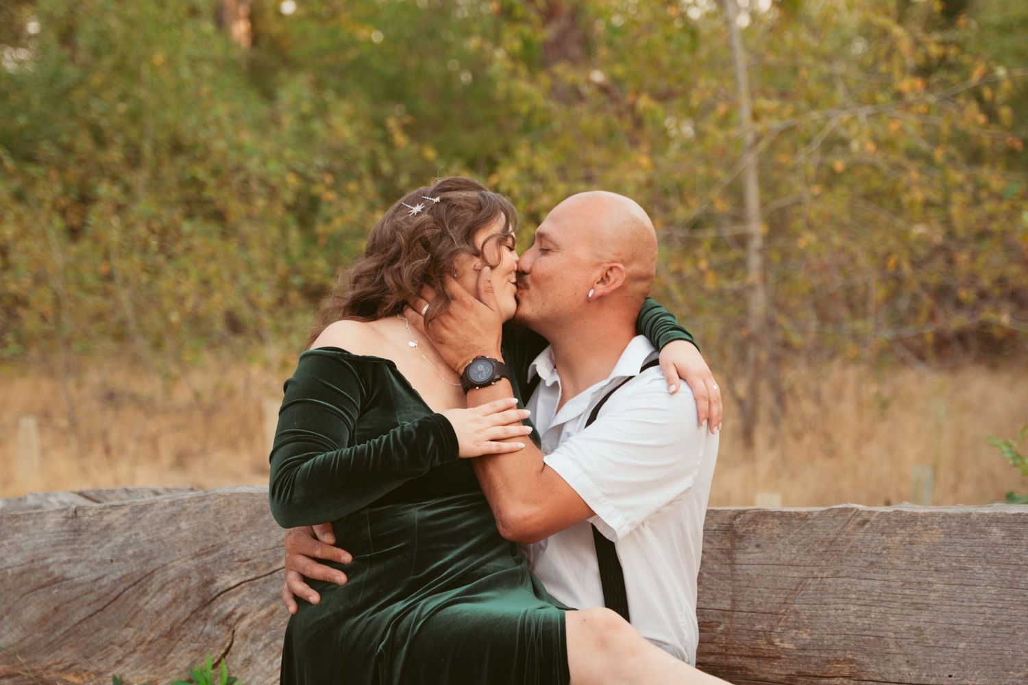 A romantic outdoor photoshoot with a couple sitting on a rustic log in a natural setting.