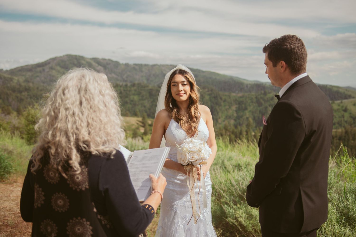 A wedding ceremony takes place on a scenic mountaintop with mountains and forests in the background.