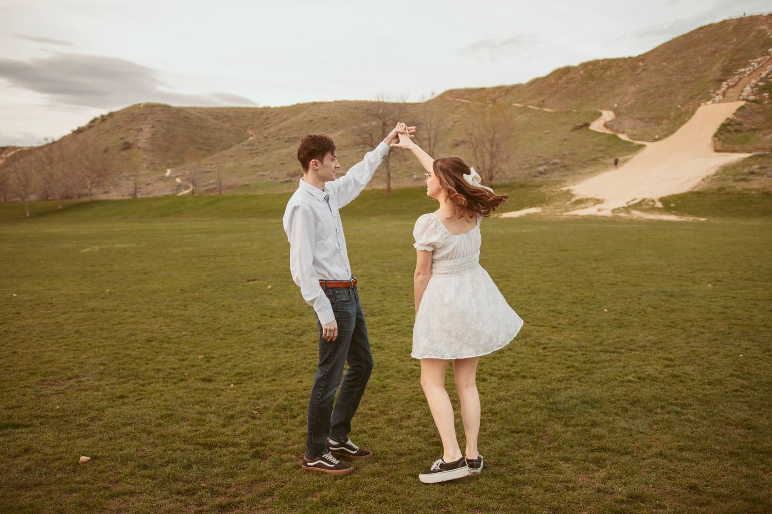 A couple dances and plays in a grassy field with mountains in the background during golden hour.