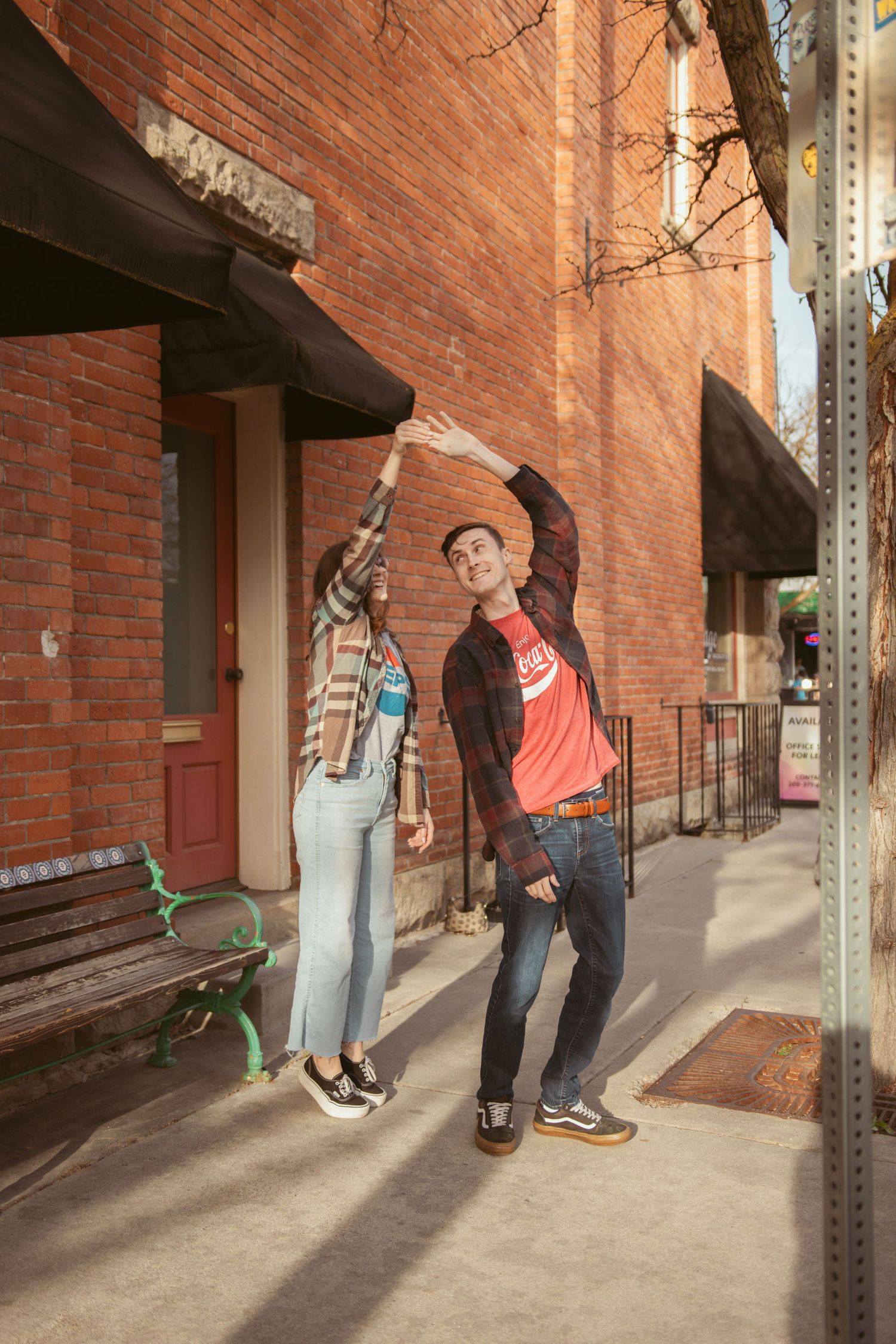 Two people strike playful poses against a brick wall on a sunny urban sidewalk.