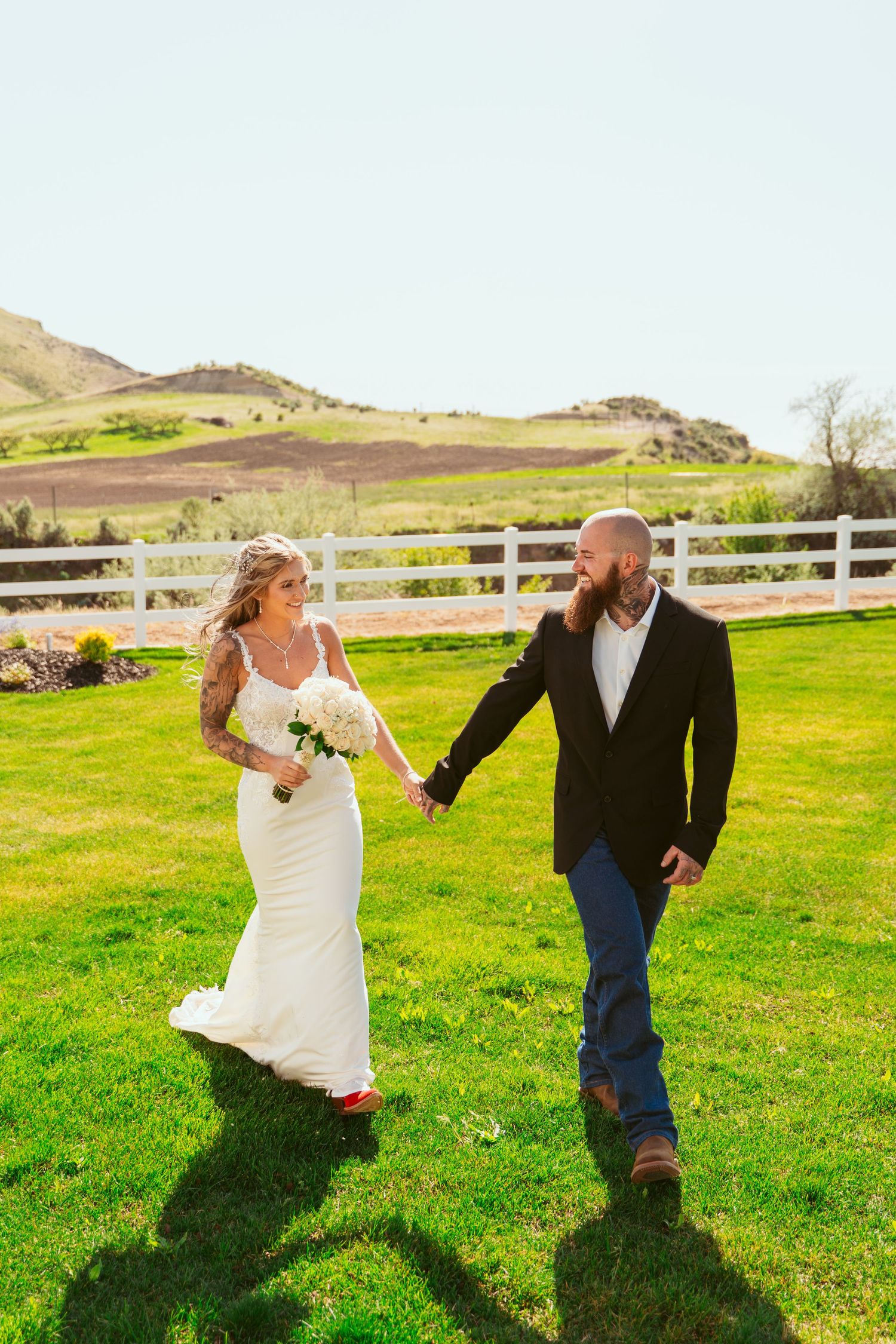 A playful wedding couple runs and dances together on a green lawn with mountains in the background.