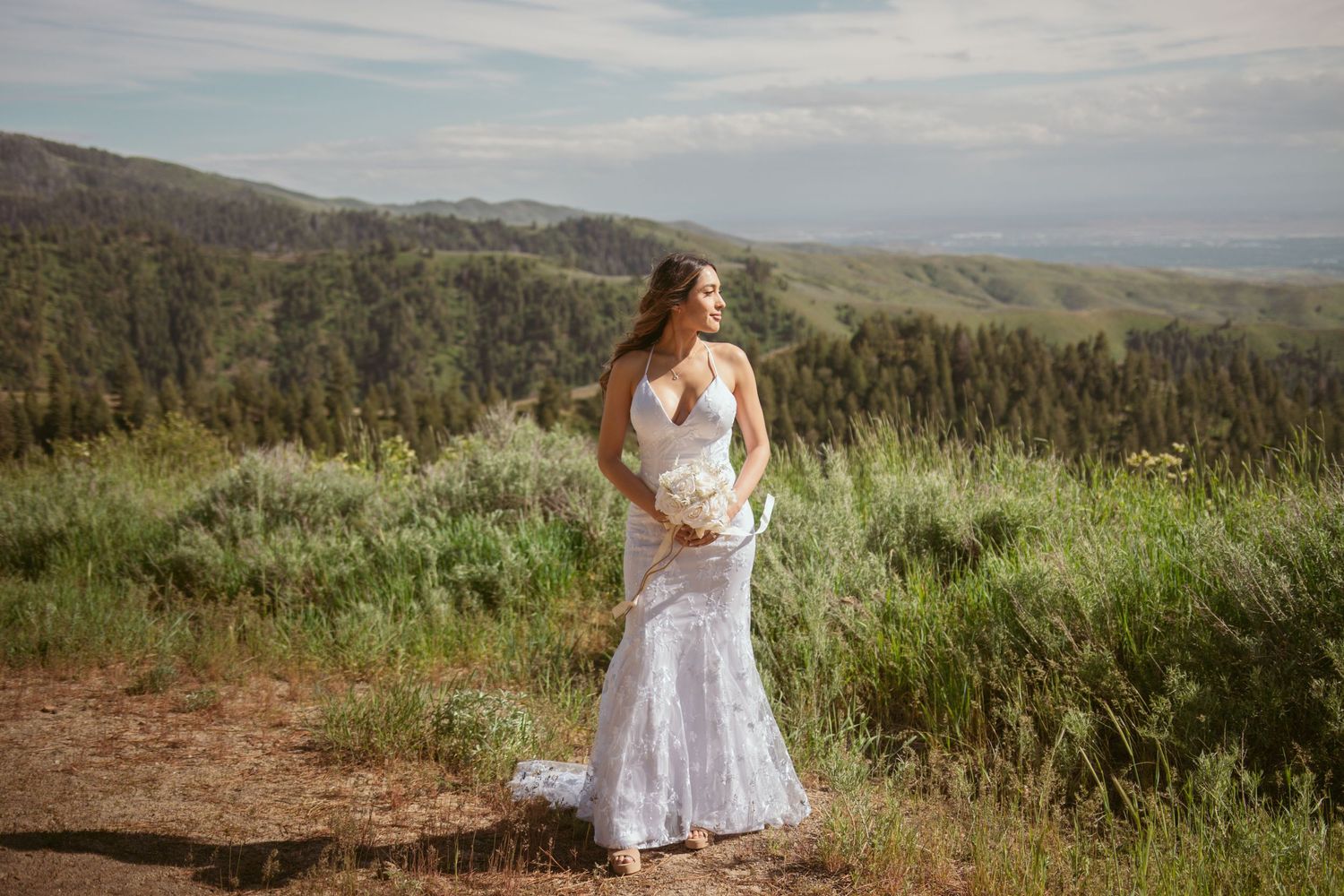 A bride in a lace wedding dress poses on a mountain trail with scenic valley views.