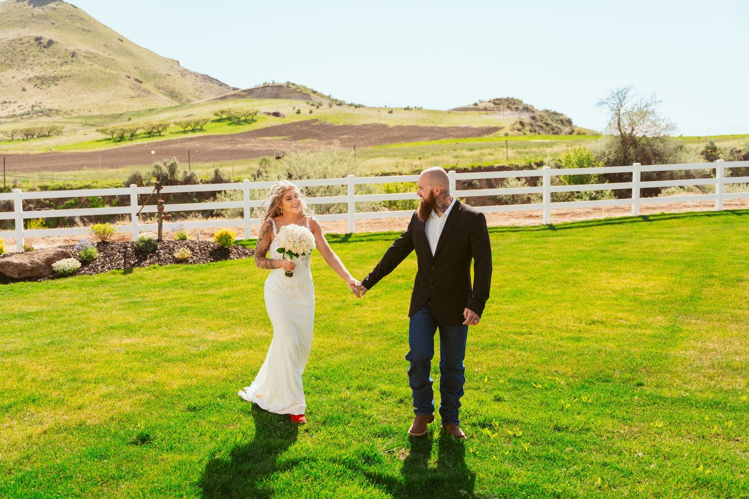 Couple strolls together on grassy field with white fence and hills in background.