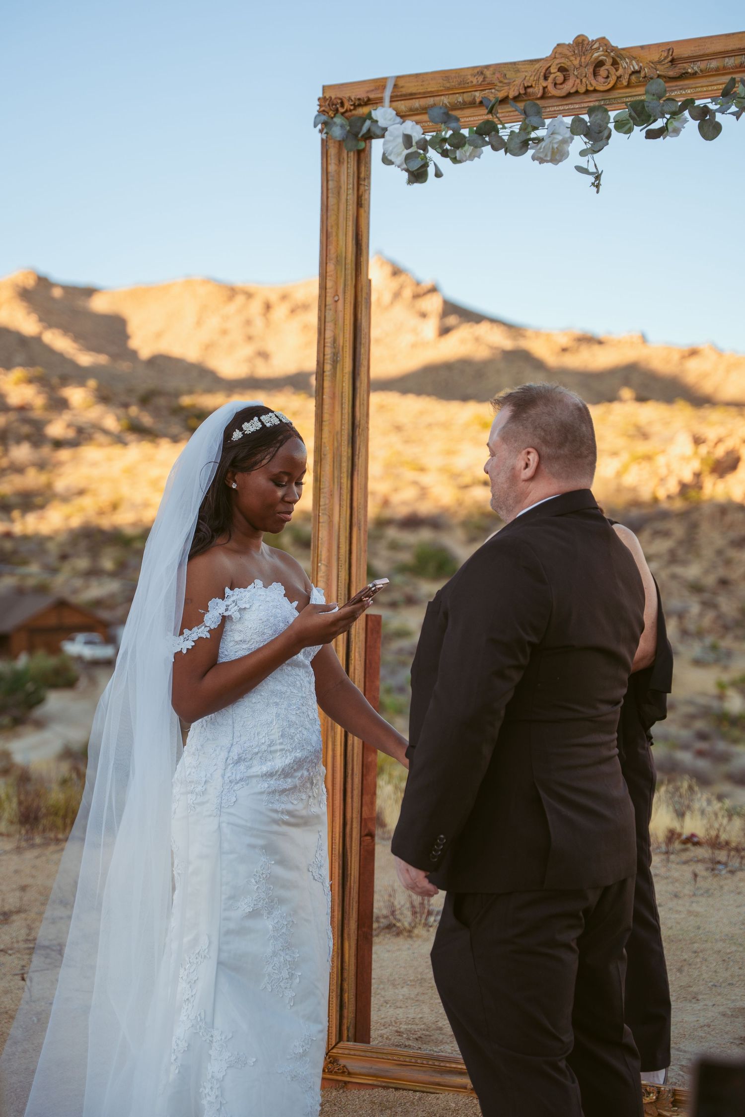 A wedding ceremony takes place in a desert landscape with a wooden arch and mountains in the background.