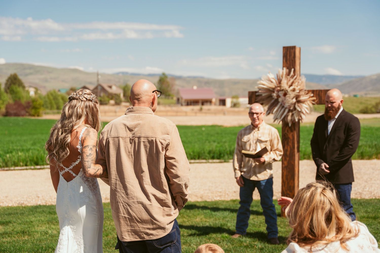 Rustic outdoor wedding ceremony with wooden cross and white chairs on green grass against mountain backdrop.