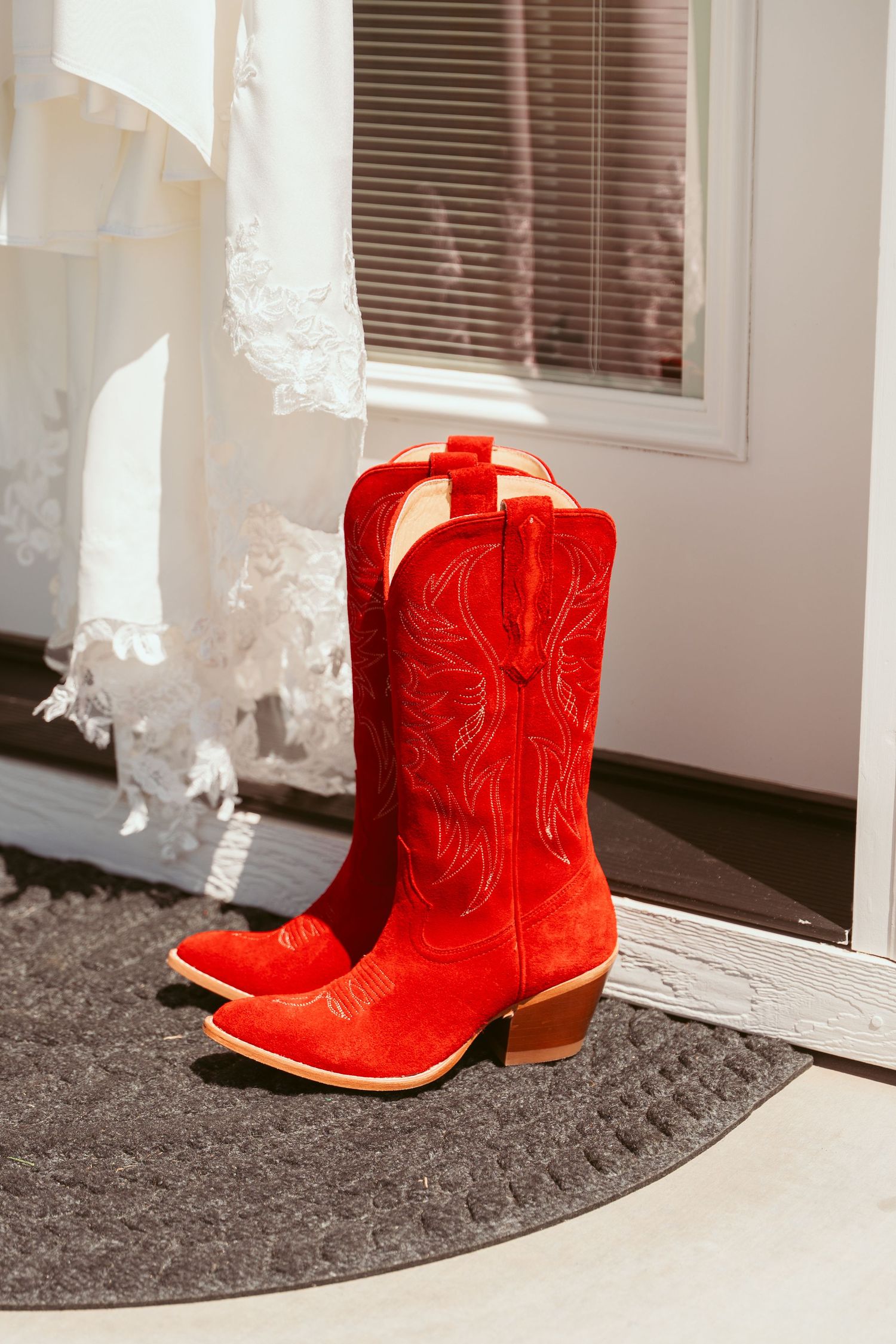 Vibrant red cowboy boots with decorative stitching on gray doormat near white dress.