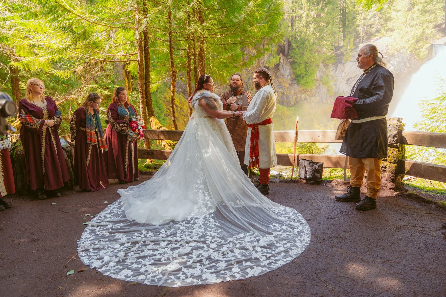 Guests gather in medieval costumes to witness a forest wedding ceremony under tall trees.