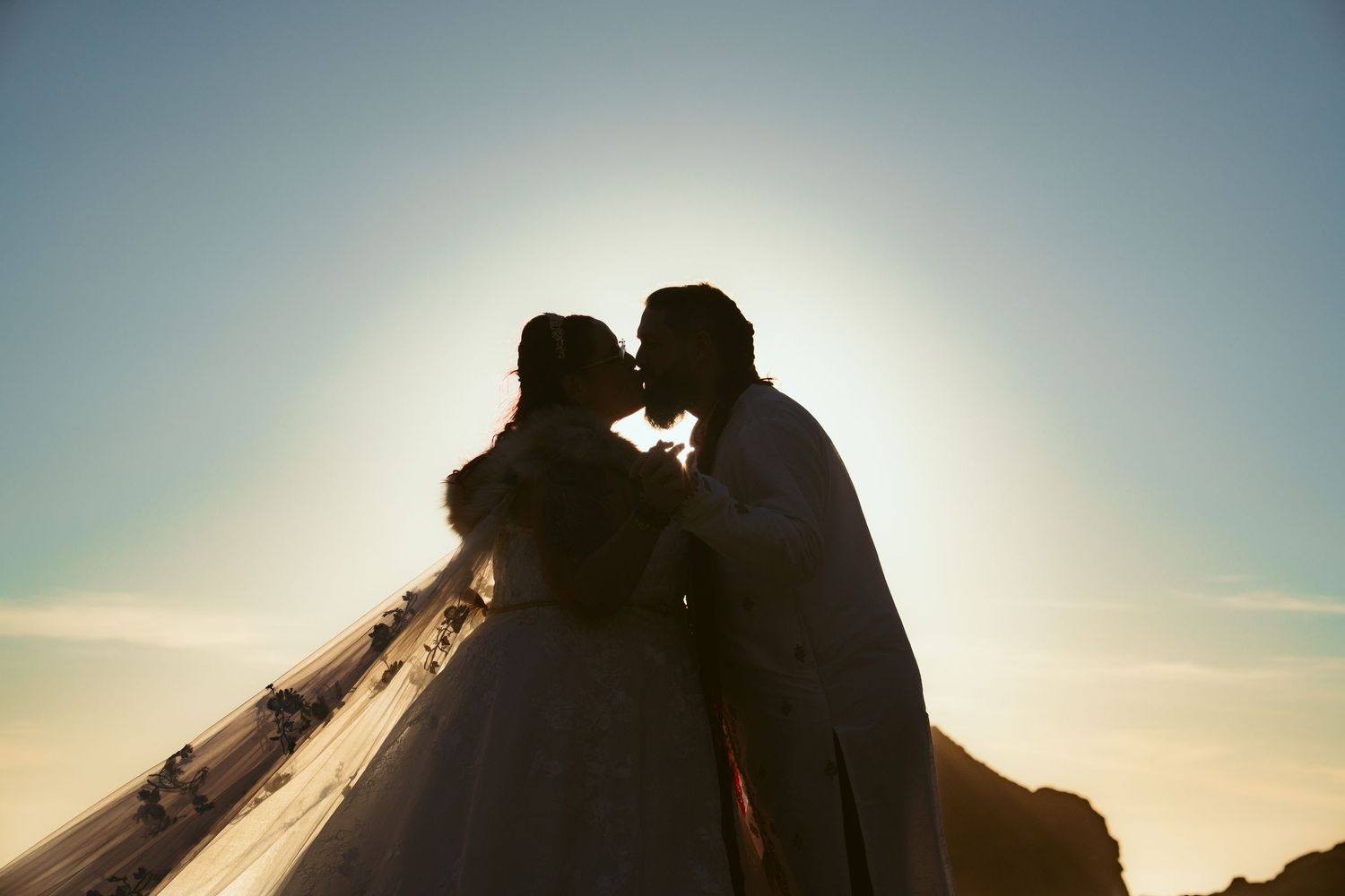 Silhouetted newlyweds share an intimate moment against a bright sunset sky.