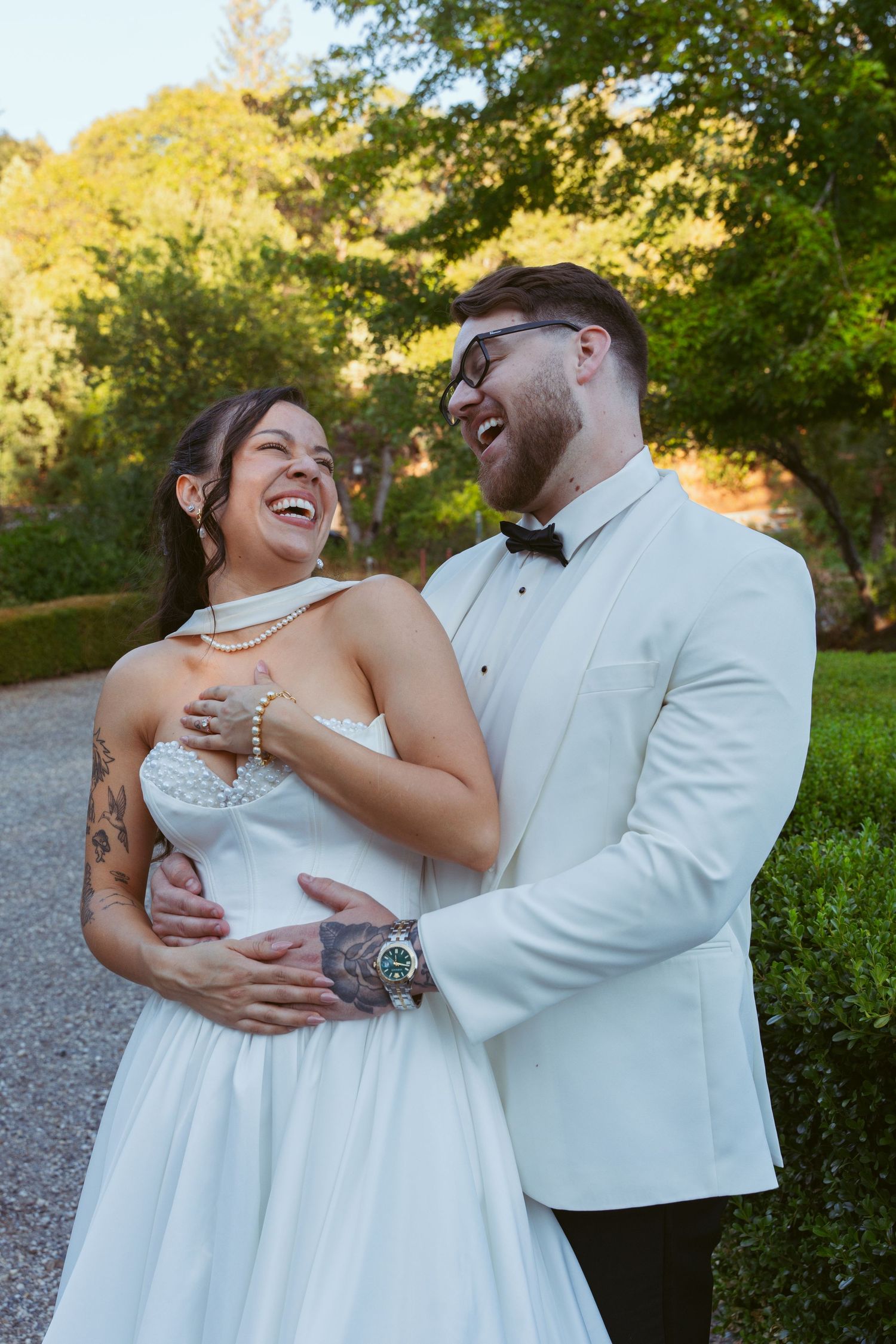 A joyful couple shares a candid laughing moment in their wedding attire against a backdrop of autumn trees.