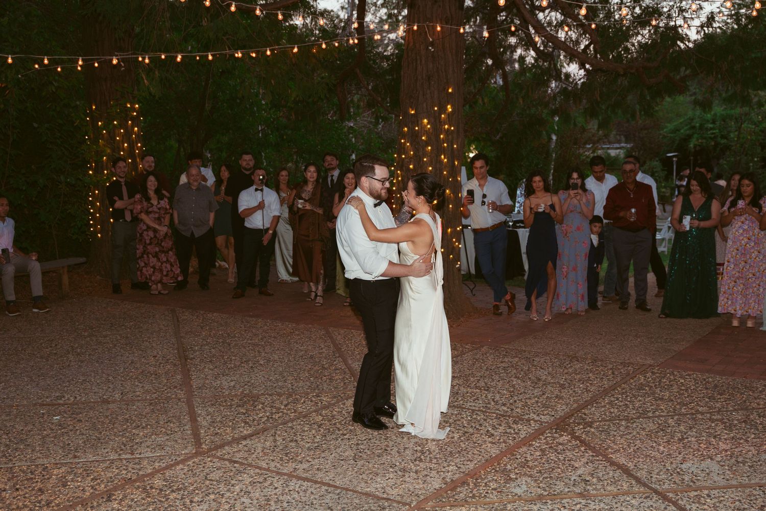 Wedding guests dancing at dusk under strung lights in a wooded outdoor venue.