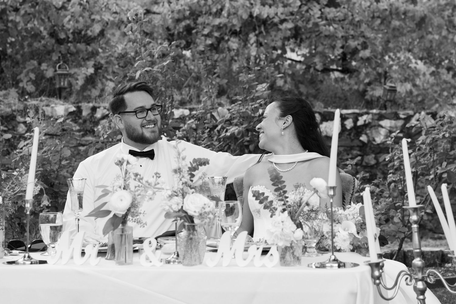 A black and white photo of a couple sharing a toast at an elegant outdoor reception table.