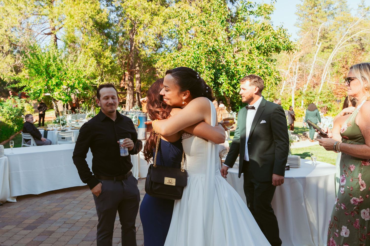 Outdoor wedding celebration with guests mingling under bright sunlight and trees.