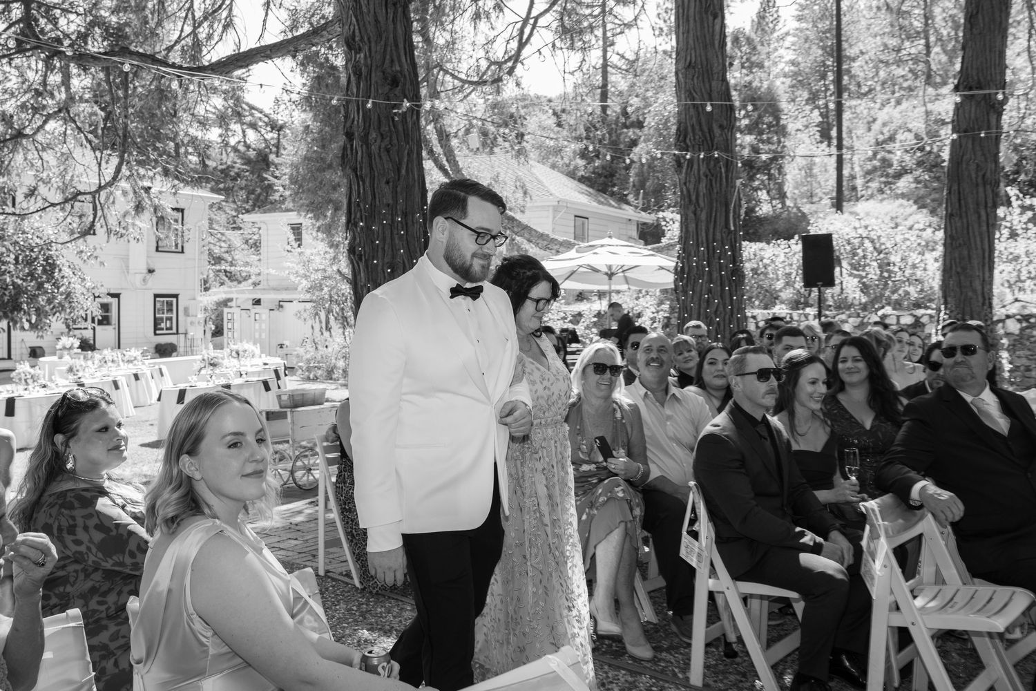 A black and white photo shows a crowd of people gathered outdoors under trees during a social event.