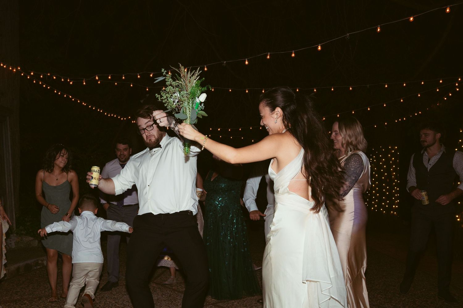Wedding celebration with guests dancing under romantic string lighting at night.