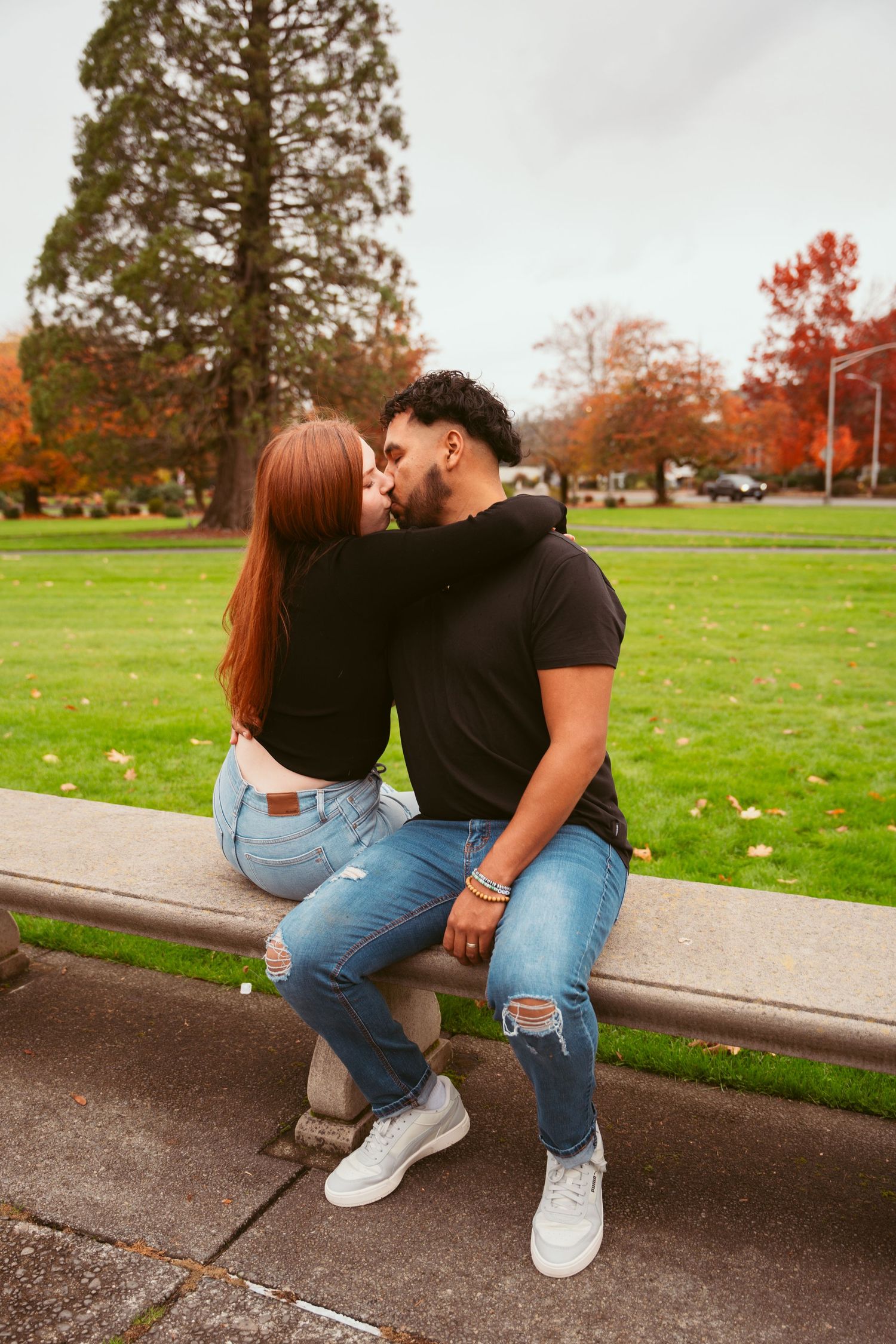 Couple sitting together on a curb with autumn trees and green grass in background.