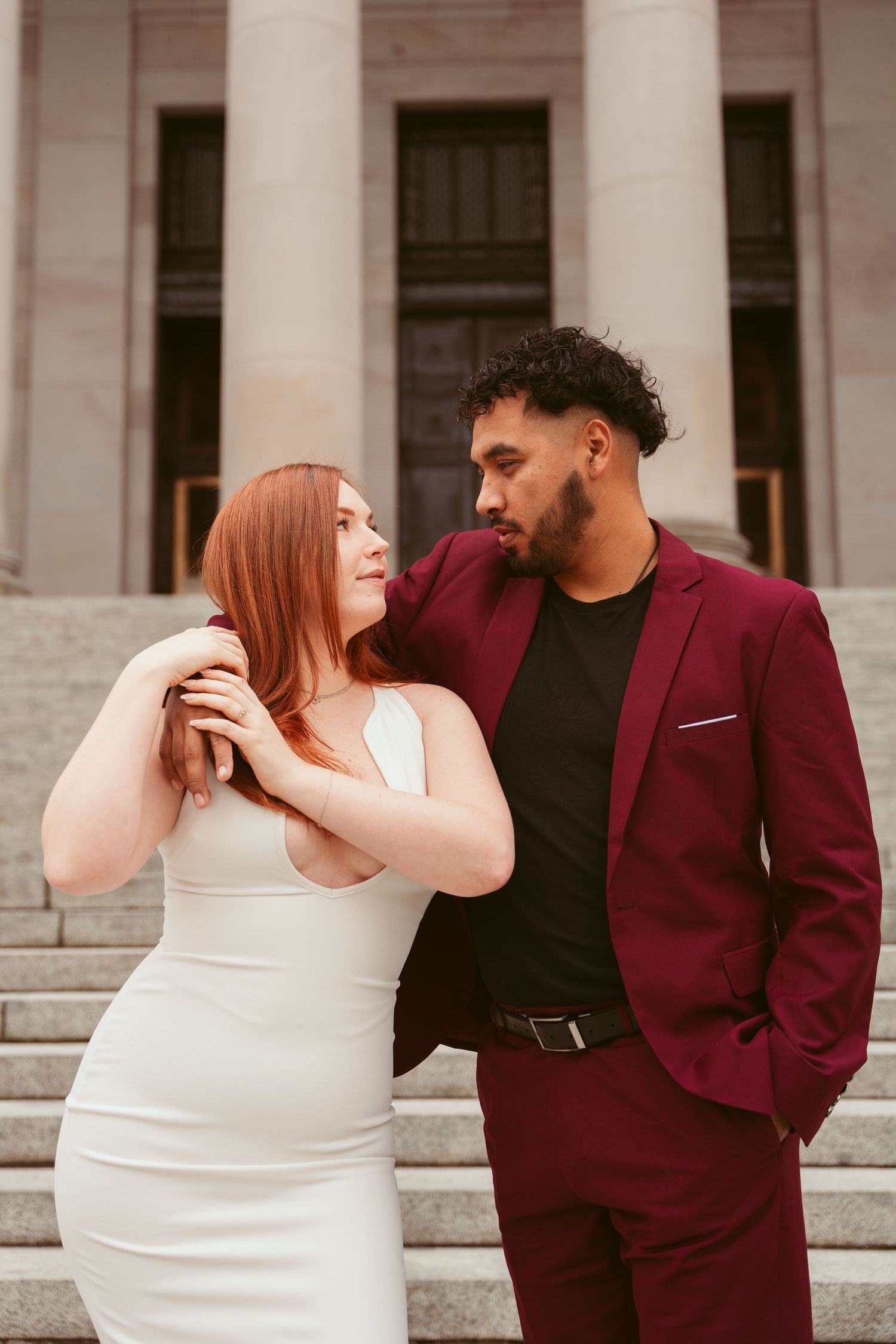 A couple in contrasting burgundy and white outfits poses affectionately on stone steps.