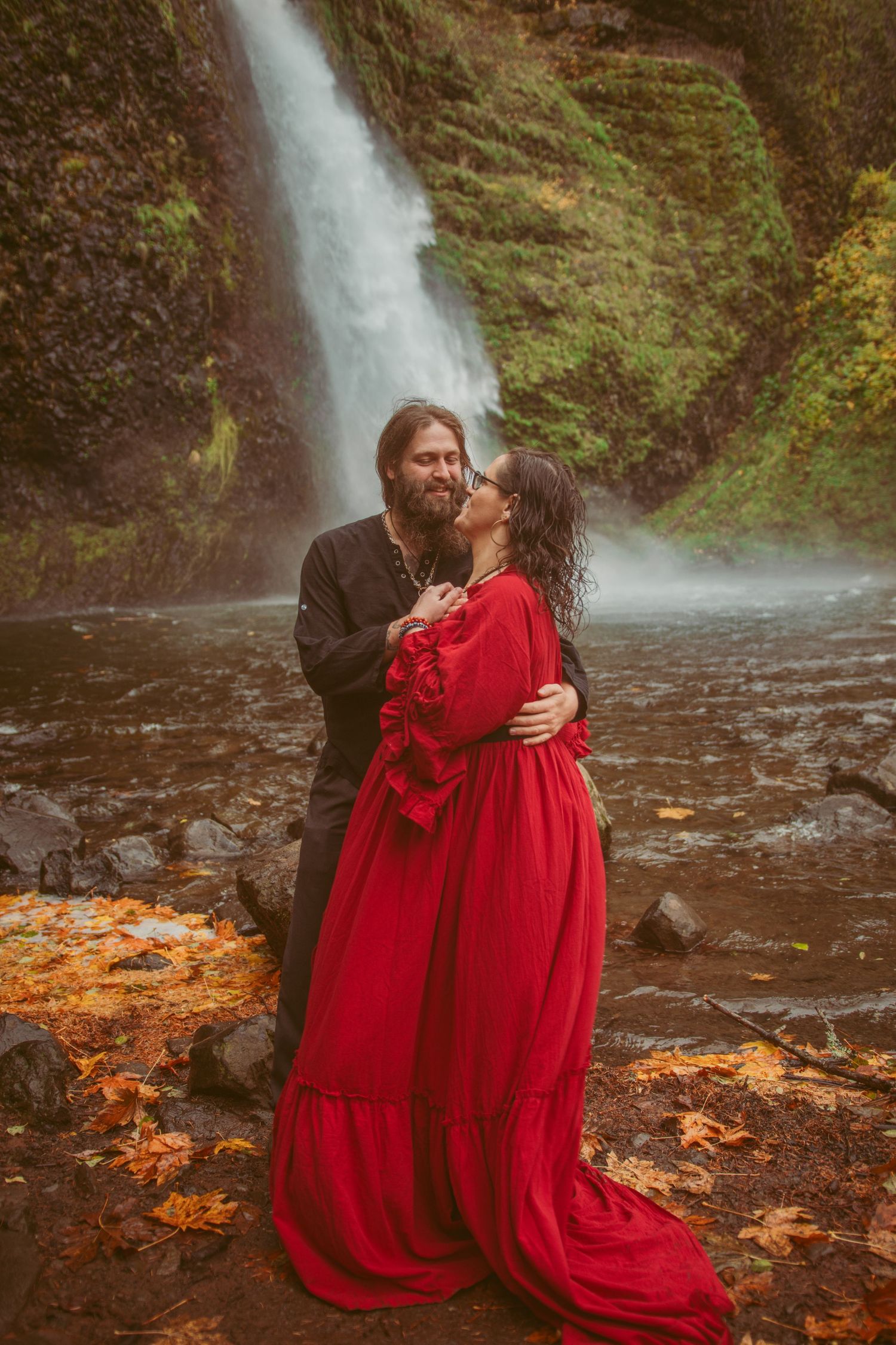 Couple embraces in front of scenic waterfall with red dress contrasting against natural backdrop.