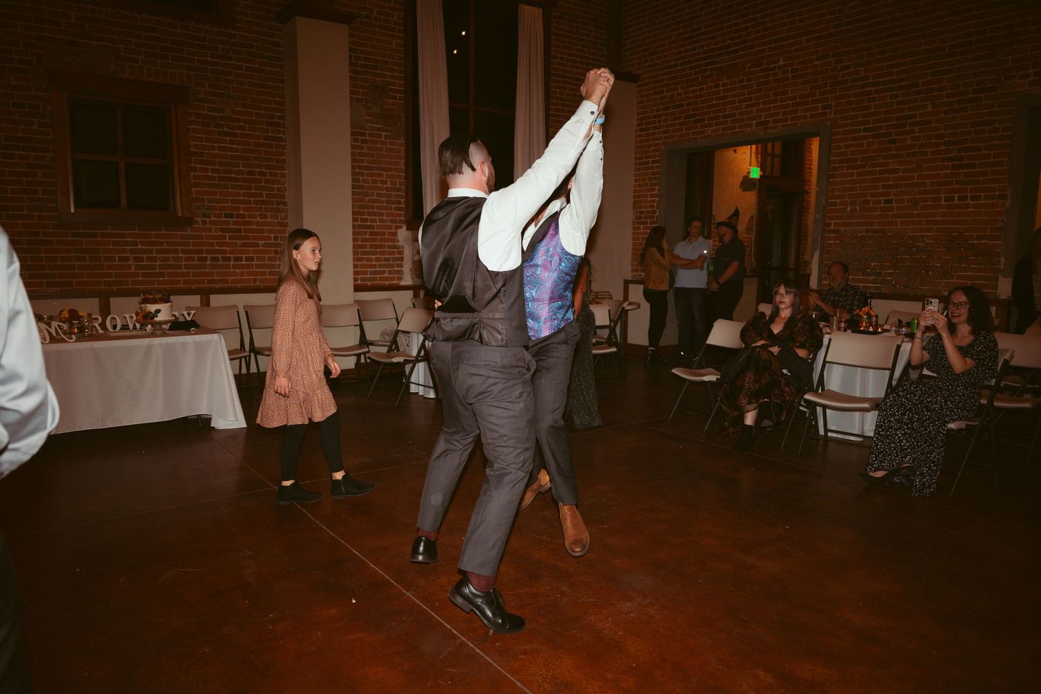 Wedding couple performing energetic dance moves on wooden floor at reception.