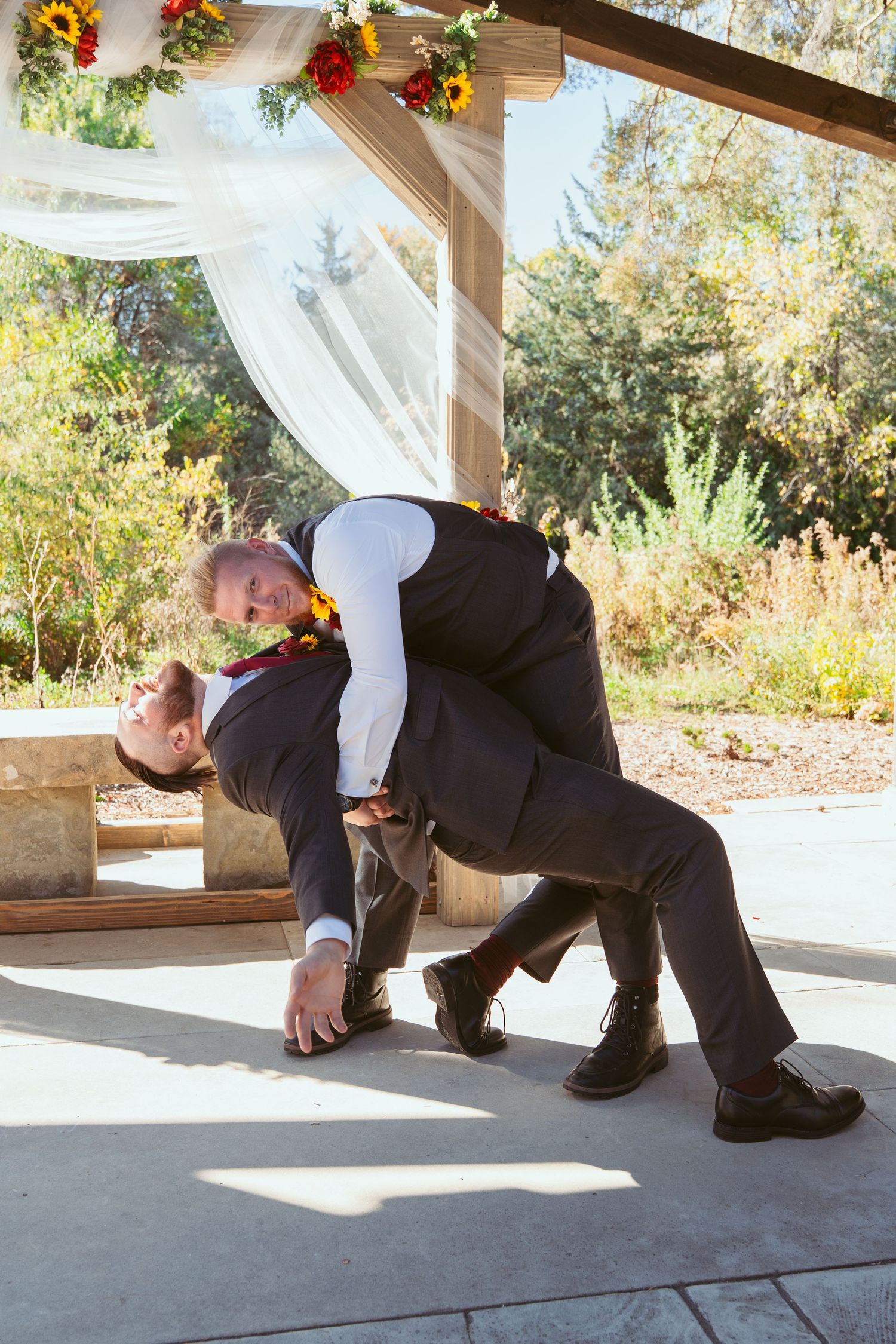 Wedding party members continue playful falling sequence under wooden pergola decorated with flowers.