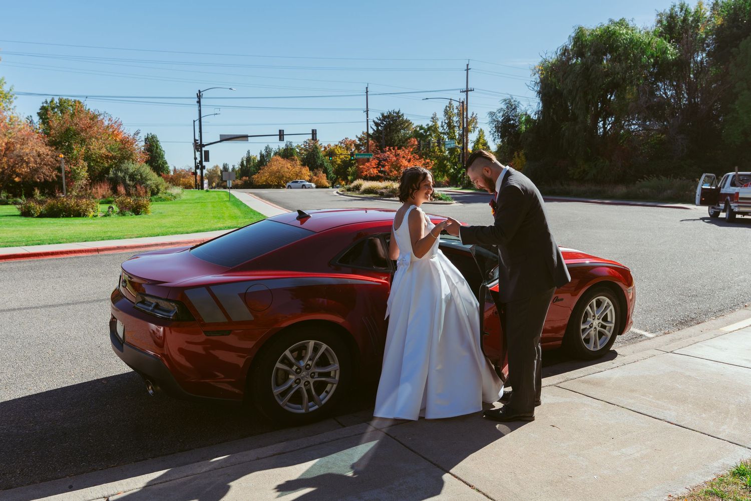 A wedding couple poses beside their red car with autumn trees in the background.