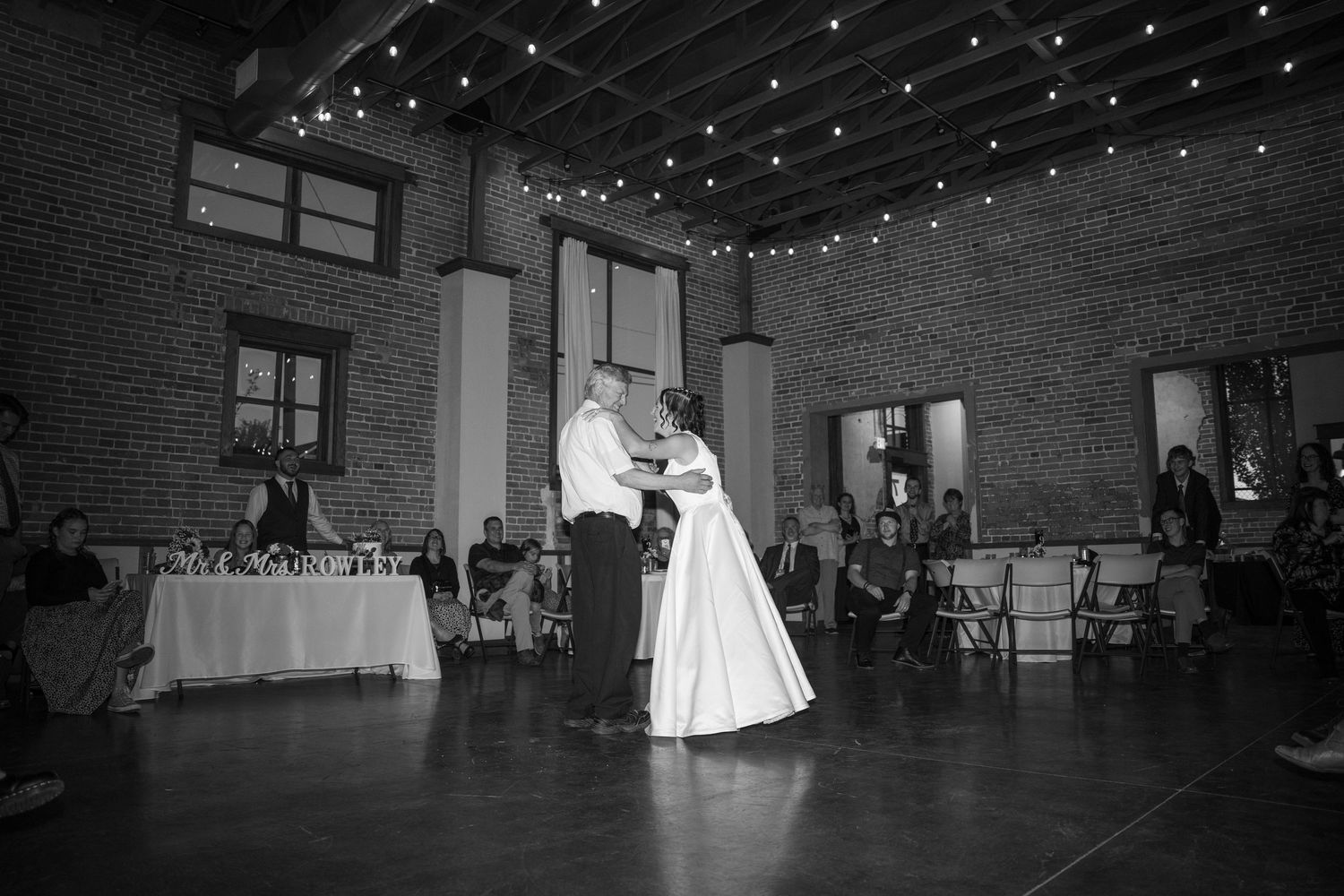Dancers move across a dark wooden floor under twinkling string lights in a brick venue.