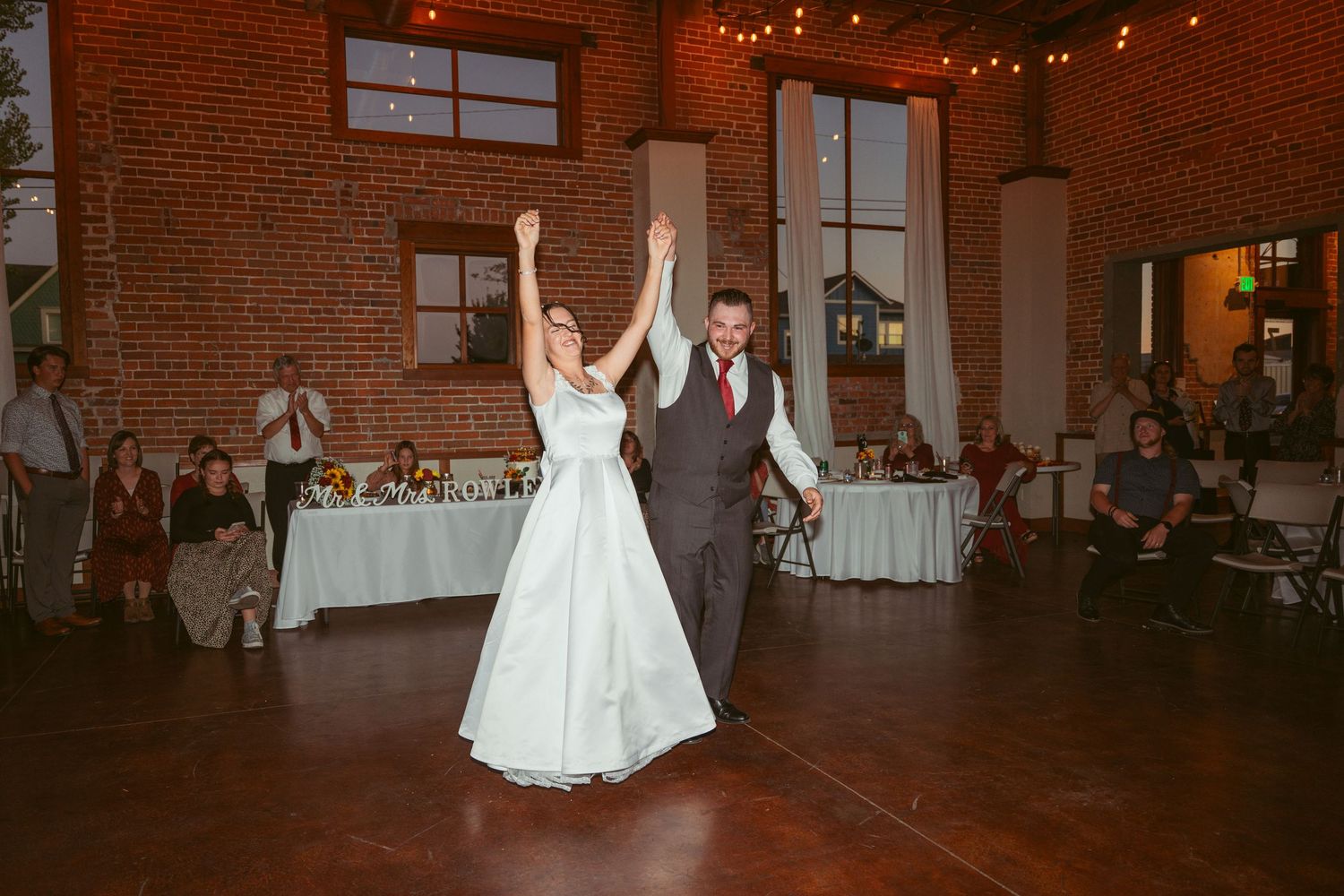 A couple shares their first dance at a wedding reception in a rustic brick venue with string lights overhead.