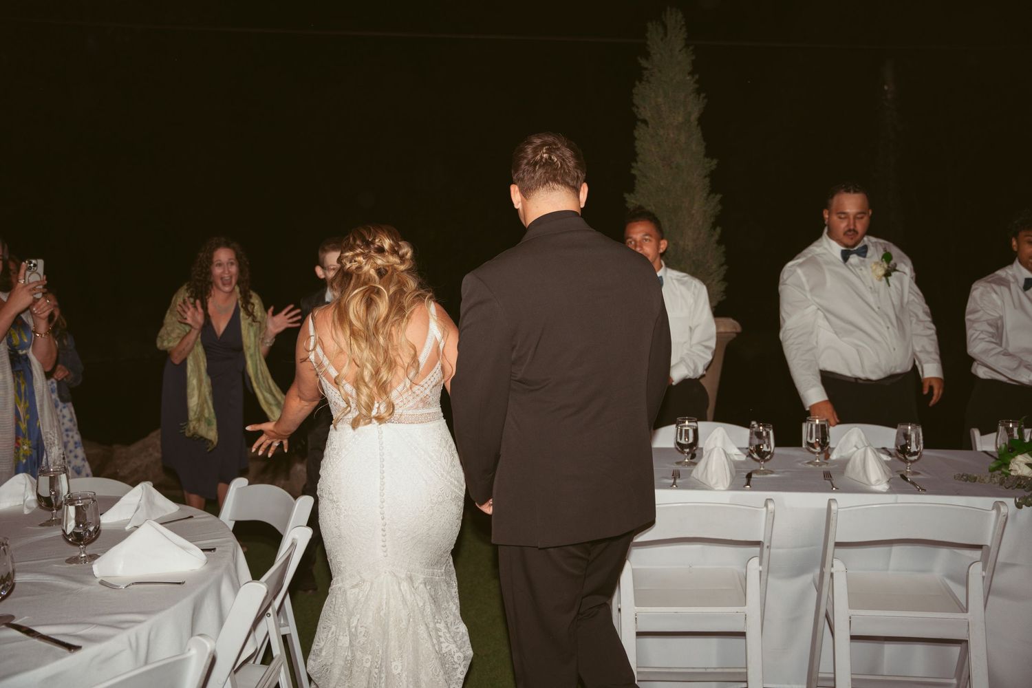 Outdoor wedding reception at night with guests seated at white tables under ambient lighting.