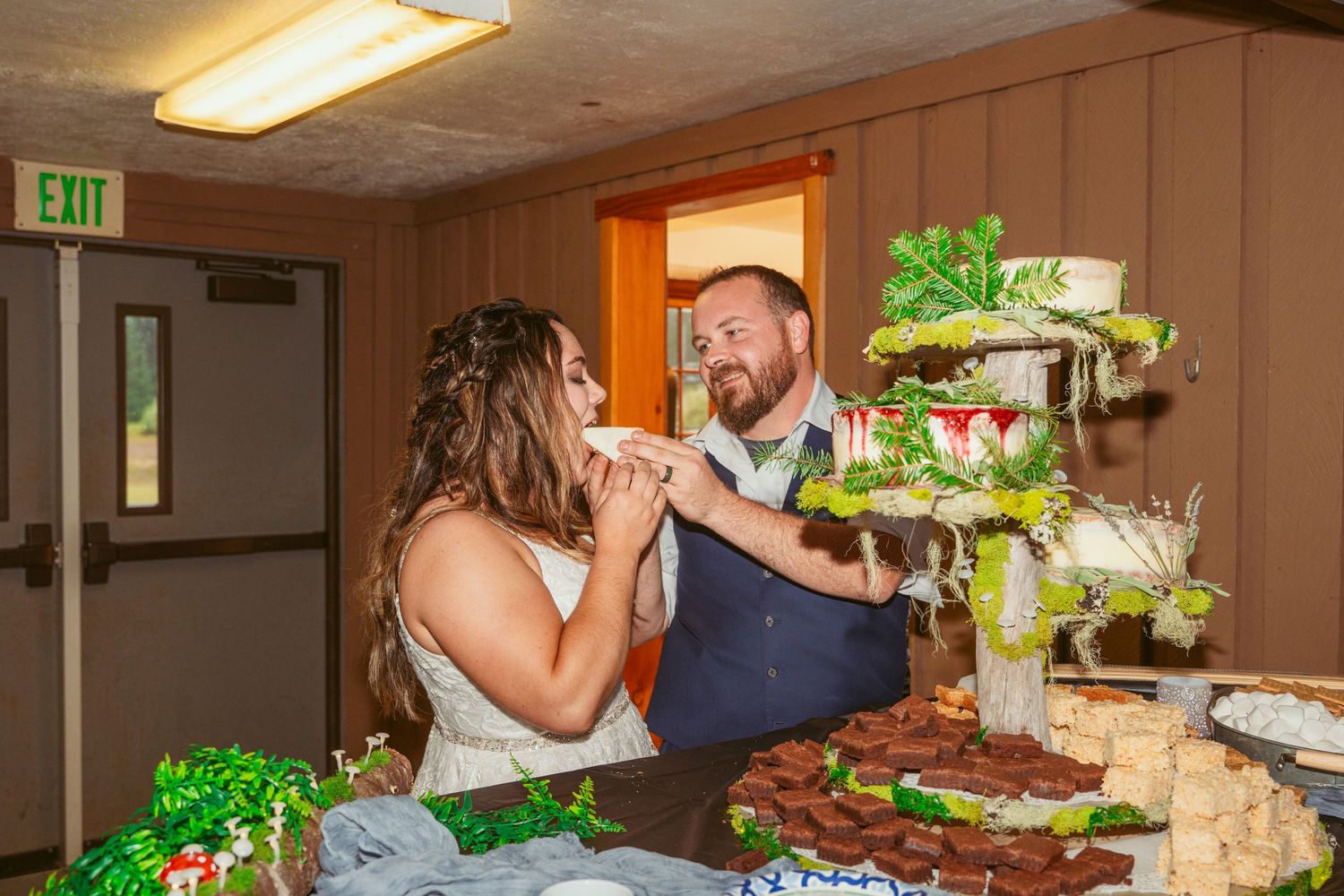 Newlyweds playfully feed each other wedding cake at their reception next to a decorated dessert table.
