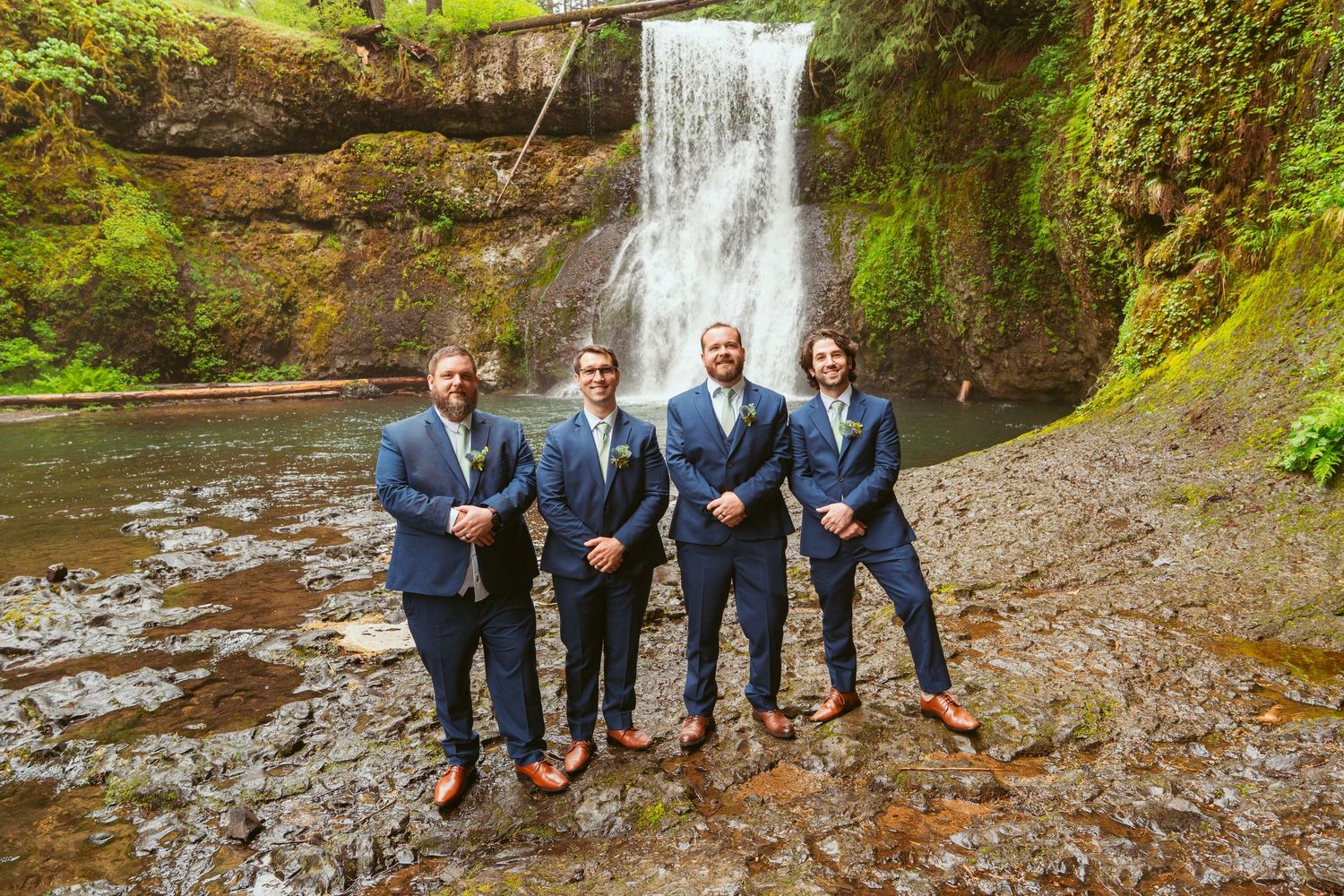 Wedding party members in navy suits pose casually by a scenic forest waterfall.